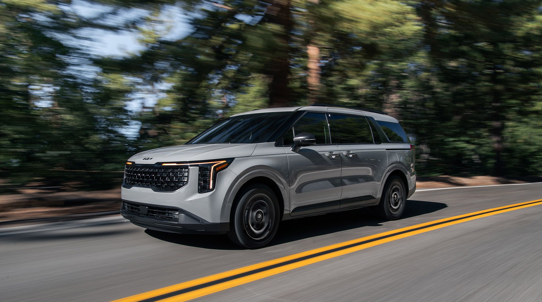 2027 Kia Carnival MPV Hybrid in silver driving along a scenic road surrounded by trees, showcasing its bold front grille, LED headlights, and sleek modern design.