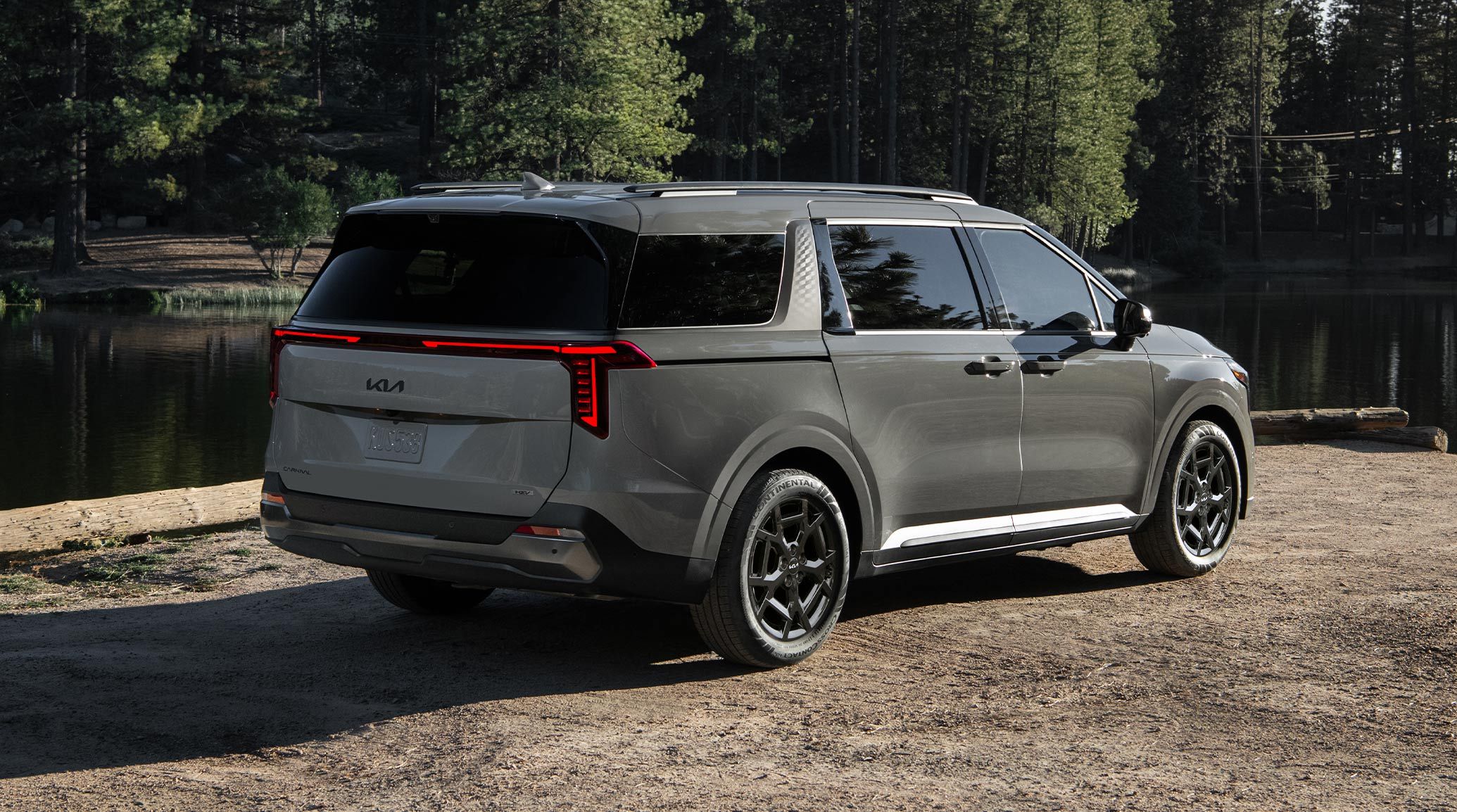 The 2027 Kia Carnival MPV Hybrid in silver is shown from the rear, parked outdoors against trees and a lake. It highlights its black wheels and refined styling.