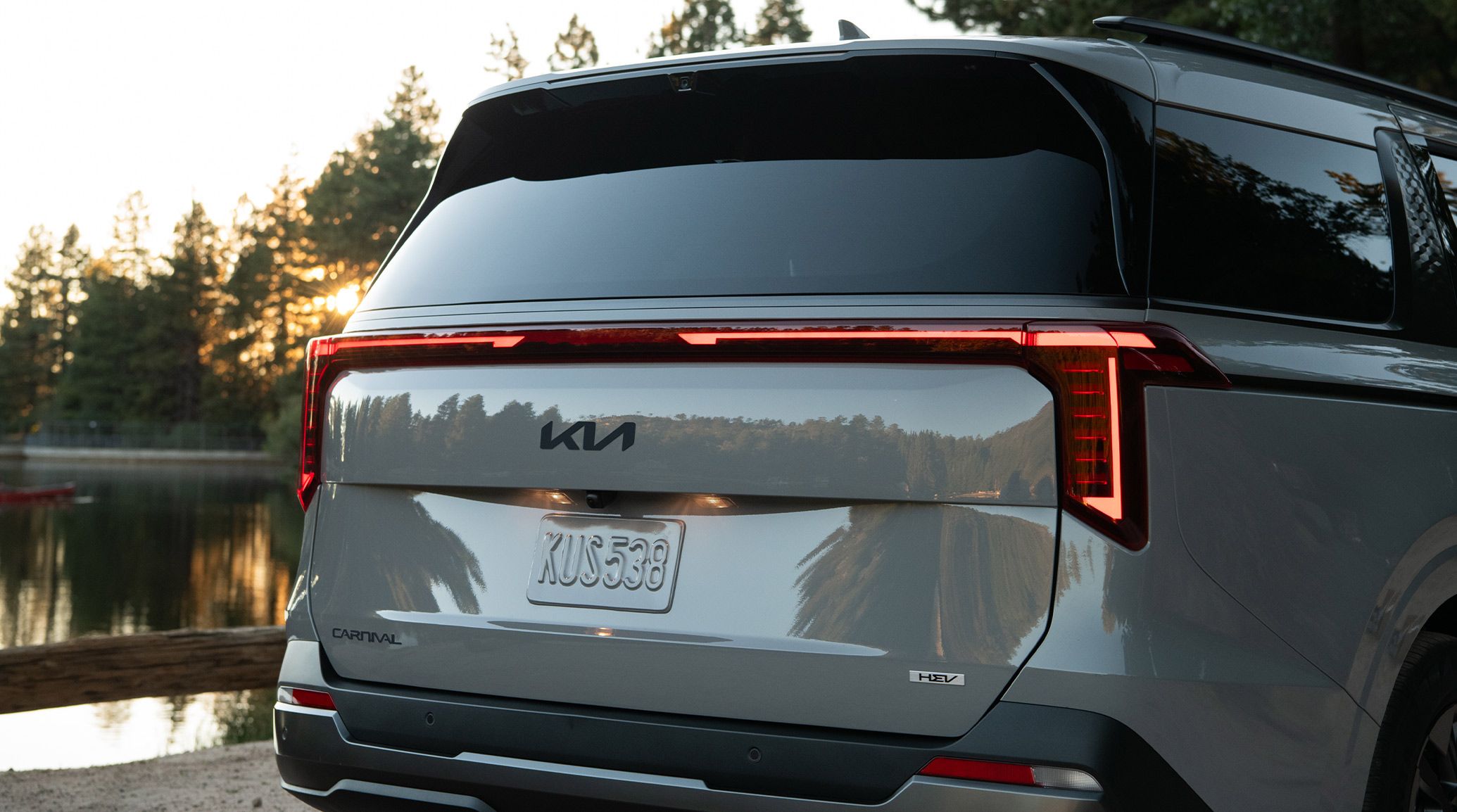A close-up rear image of the 2027 Kia Carnival MPV Hybrid, SUV-inspired minivan, near a lake at sunset, highlighting its full-width LED taillights and modern design.