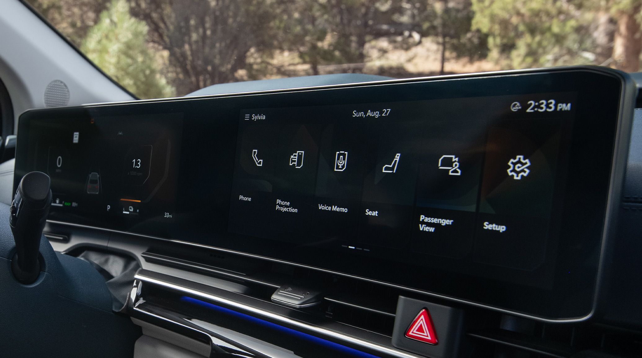 A close-up shot of the 2027 Kia Carnival MPV Hybrid interior features a wide digital display in black with navigation and vehicle controls integrated into a sleek dashboard layout, with trees peeking out through the windshield.
