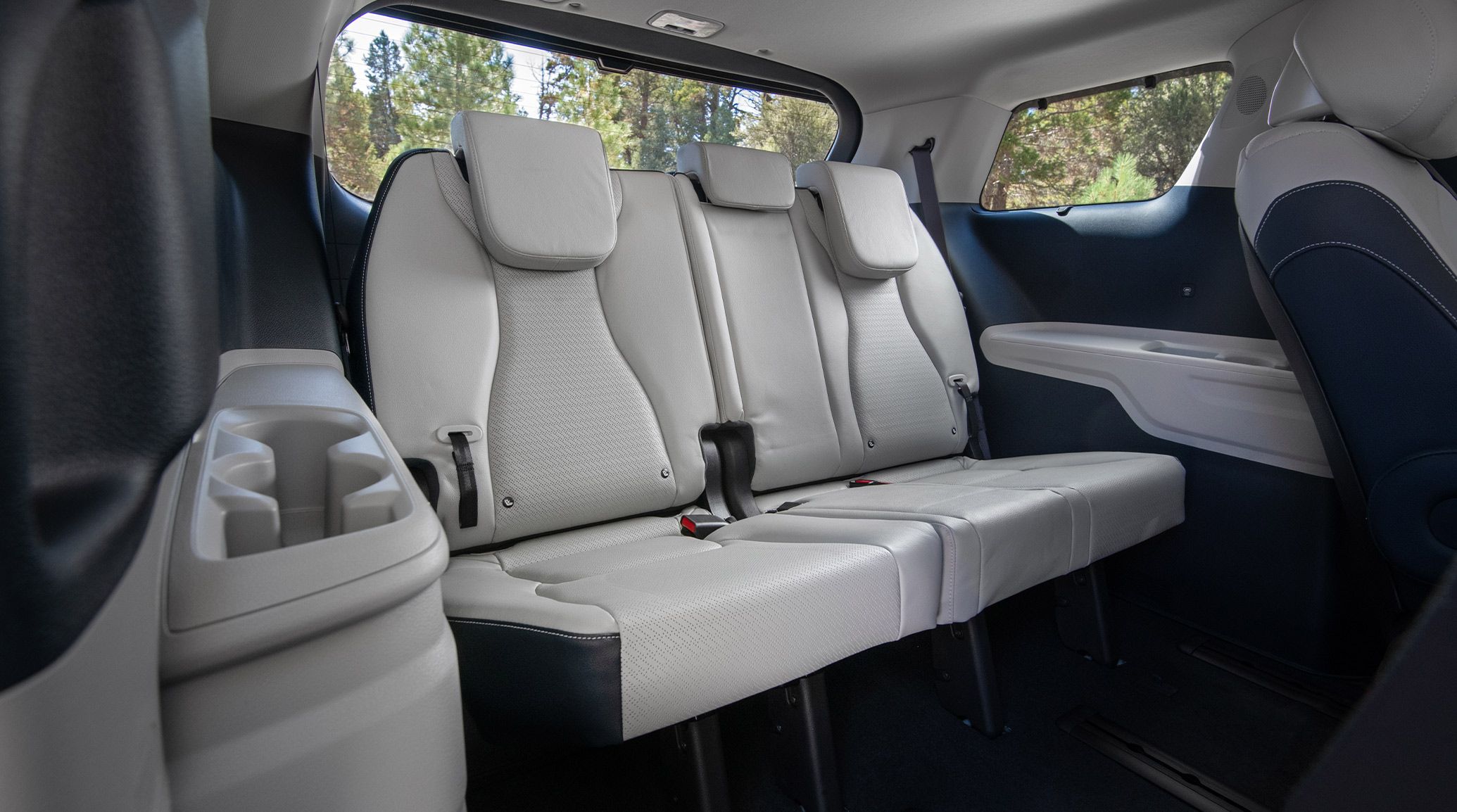 An image highlighting the gray third-row seating area of the 2027 Kia Carnival Hybrid. The image highlights the back row seating that has a 60/40 split-folding and the spacious cupholders, with trees visible through the rear-view window.