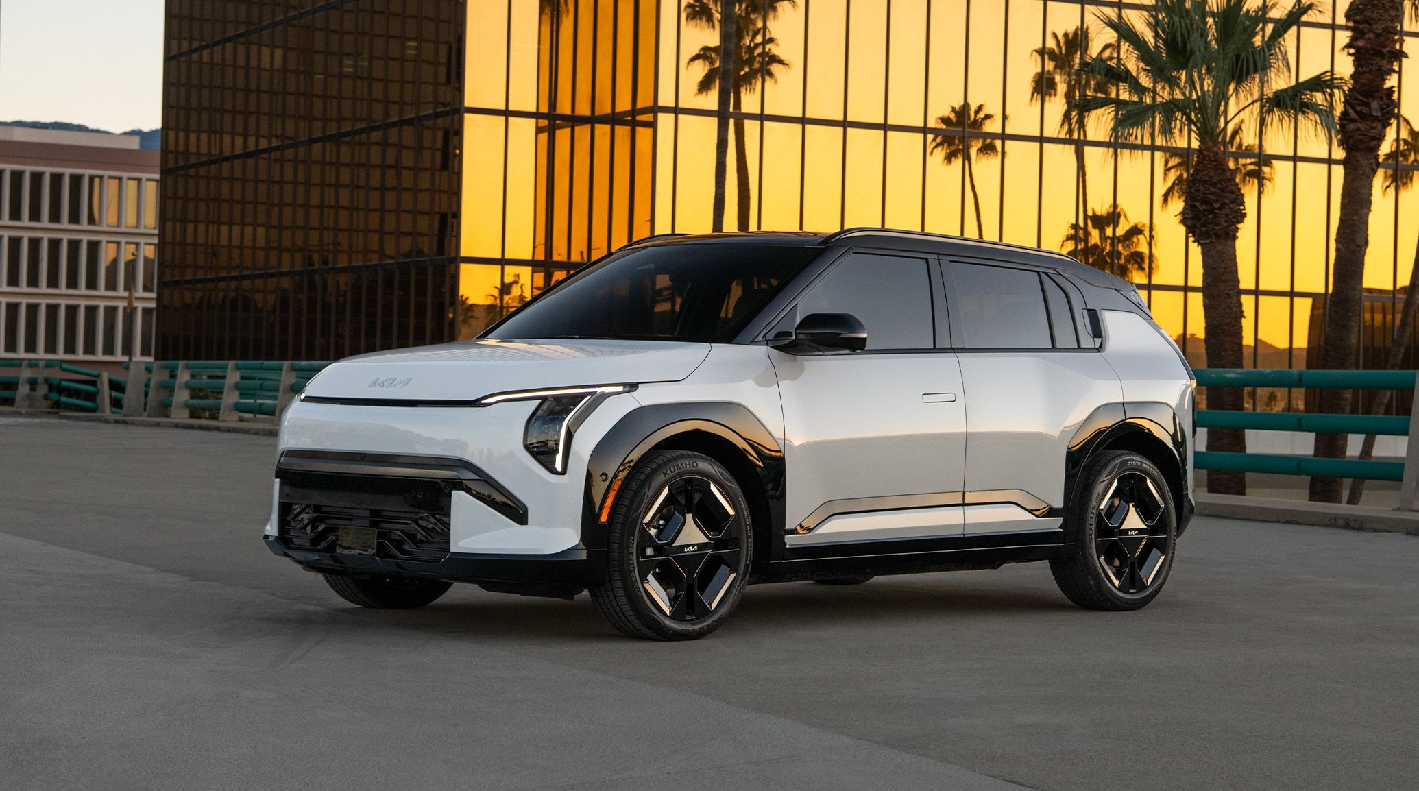 Front side angle photograph of the 2027 Kia EV3 electric compact SUV, showing silhouette, tires, headlights, door handles, and other exterior features of the electric vehicle. The EV3 is parked on asphalt with a golden glass skyscraper and palm trees in the background.