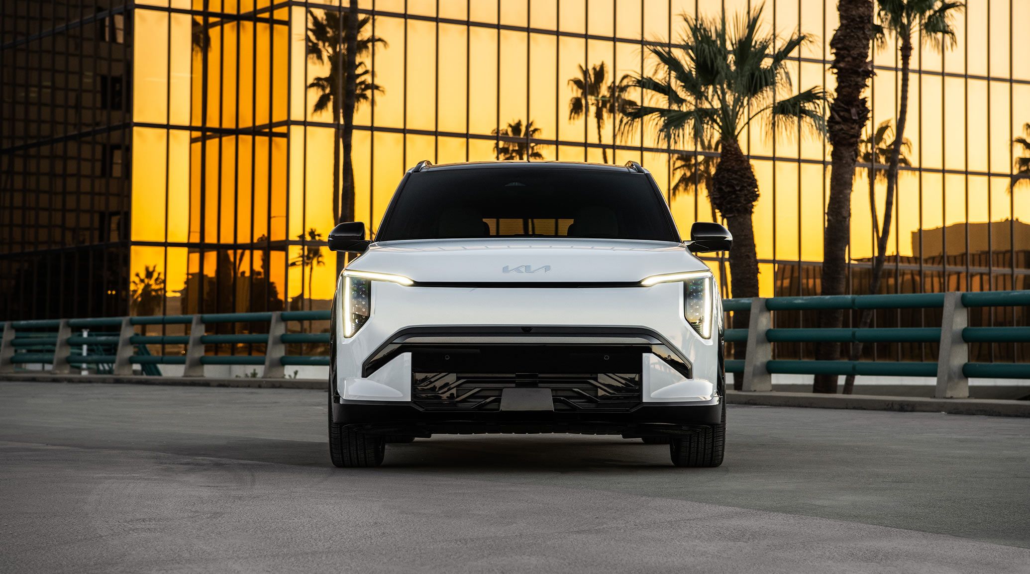 Straight-on front view of the 2027 Kia EV3 compact electric SUV, highlighting its headlights, grille, and overall shape. It is parked on asphalt with a golden skyscraper and palm trees behind it.