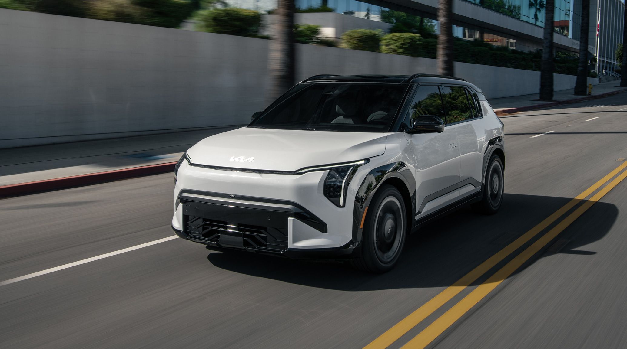 Dynamic shot of the 2027 Kia EV3 compact electric SUV in white driving rapidly down an urban road, with motion-blurred buildings and palm trees in the background. This photograph is meant to portray the excitement and fun of driving the EV3.