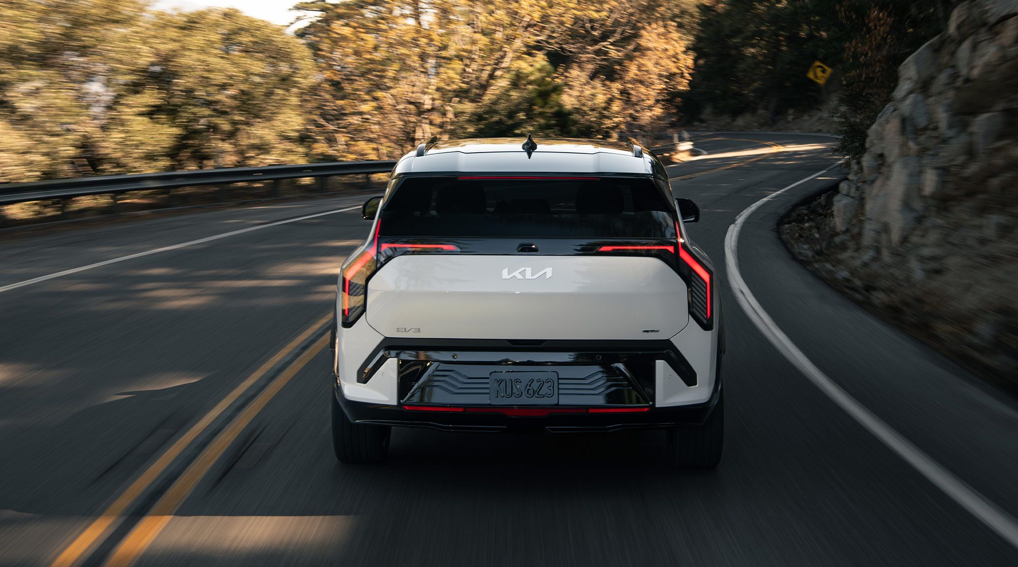 Straight-on photograph of the rear of the 2027 Kia EV3 compact electric SUV showcasing the back windshield, closed trunk door, and license plate. The EV is driving through a twisting road surrounded by rocks and trees, with sun and shadows across the asphalt.