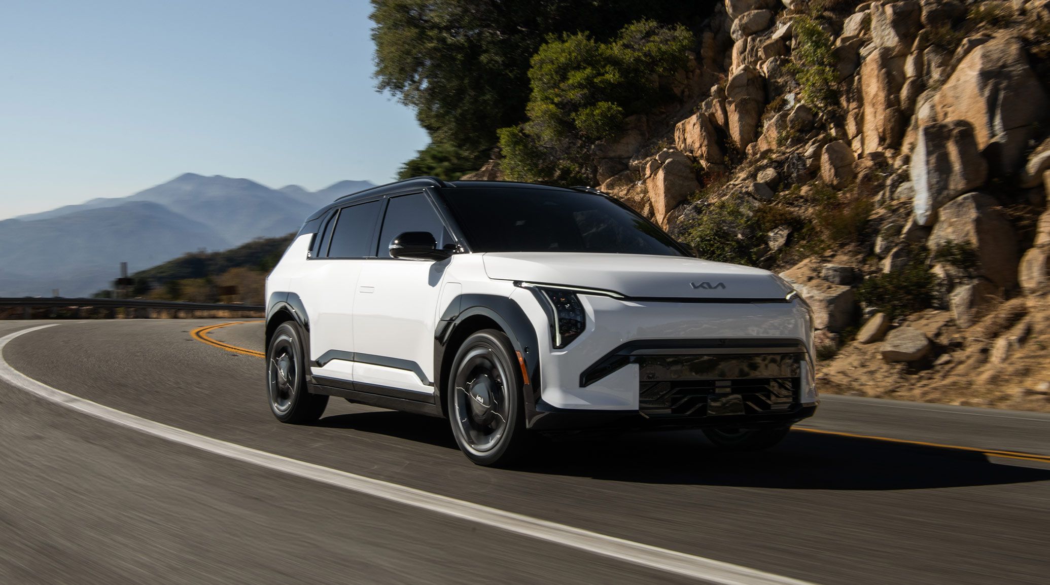 Front angle view of the 2027 Kia EV3 compact electric SUV as it turns a corner on a curvy road with rocky mountains and trees in the background. The front grille and Kia logo are visible as well as the black highlighting on white.
