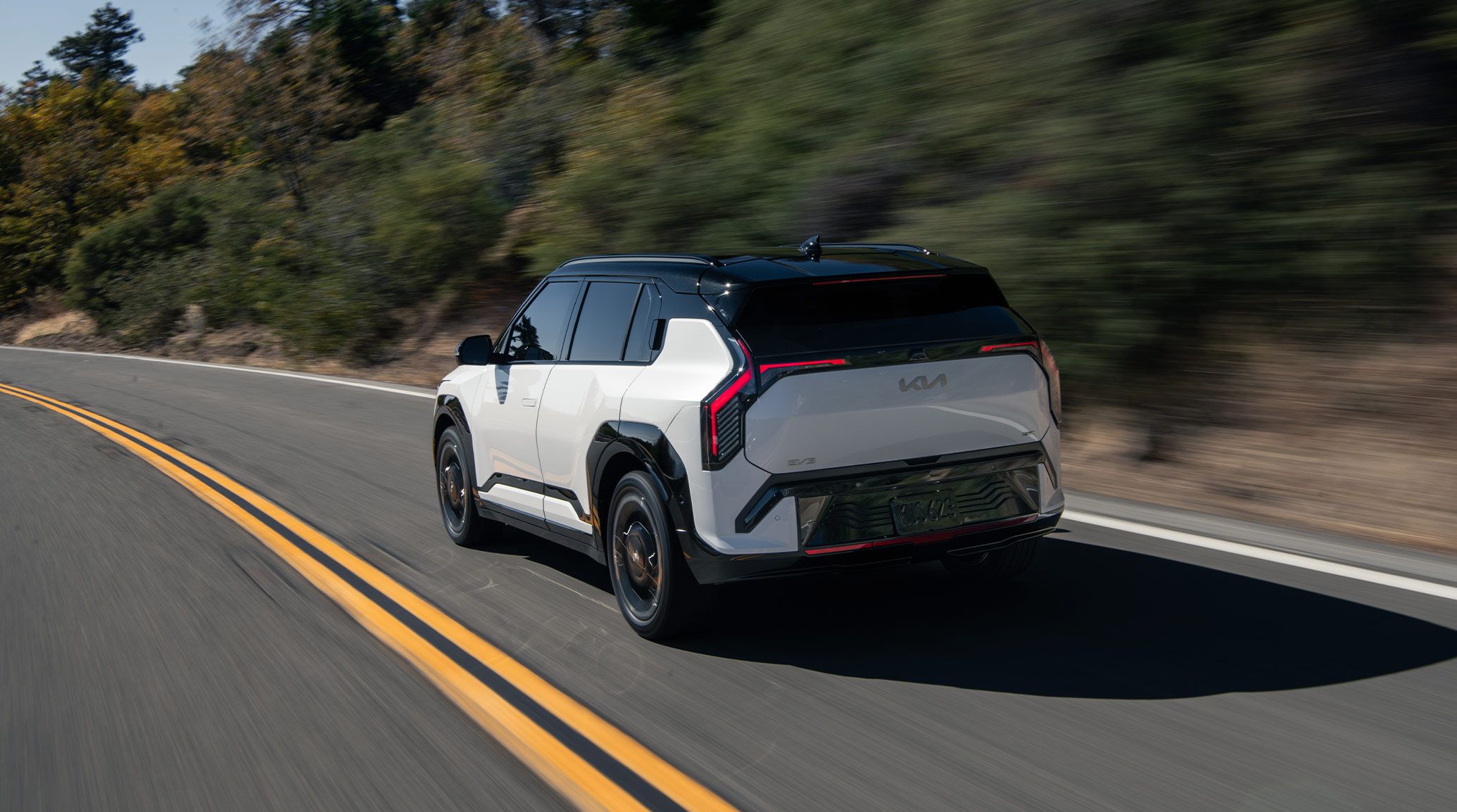 Rear side angle of the 2027 Kia EV3 compact electric SUV in white with black highlighting as it rounds a wide curve in a rural, forest setting.