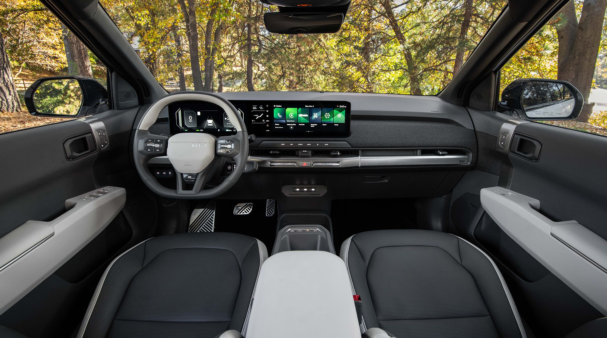 Interior wide-angle view of the 2027 Kia EV3 electric SUV showing seats, pedals, steering wheel, digital display, and more, with trees visible out the windows and windshield.