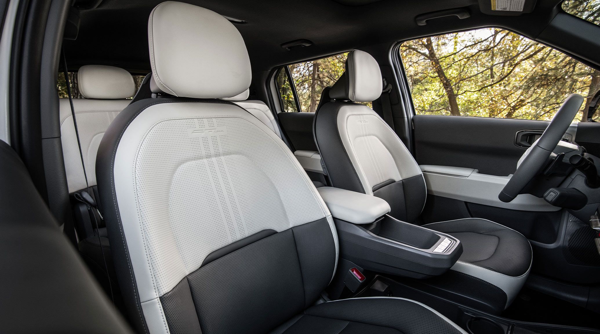 Interior angle view of the 2027 Kia EV3 compact SUV, showcasing the modern, two-tone seats with headrests and cushioning. Trees are visible out of the side windows.