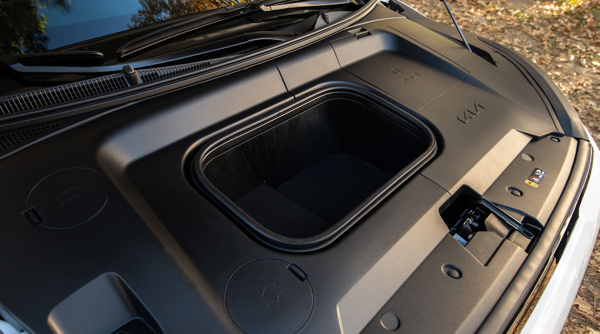 A photo of the 2027 Kia EV3 compact electric SUV with its hood lifted up and propped open, showing the front interior trunk storage space (frunk) as well as smooth surfaces and access panels.
