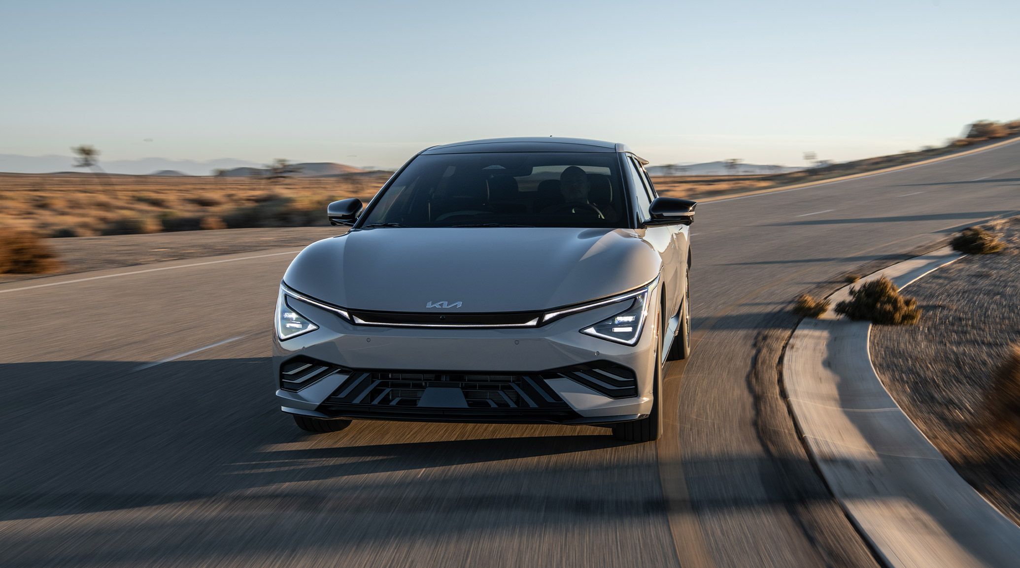 An exciting action shot of the 2026 Kia EV6 Wind AWD electric CUV in grey as it drives towards the viewer, coming down from a curvy desert road with sand and shrubs visible in the background. The CUV looks sleek and dynamic with its front design and LED headlights.