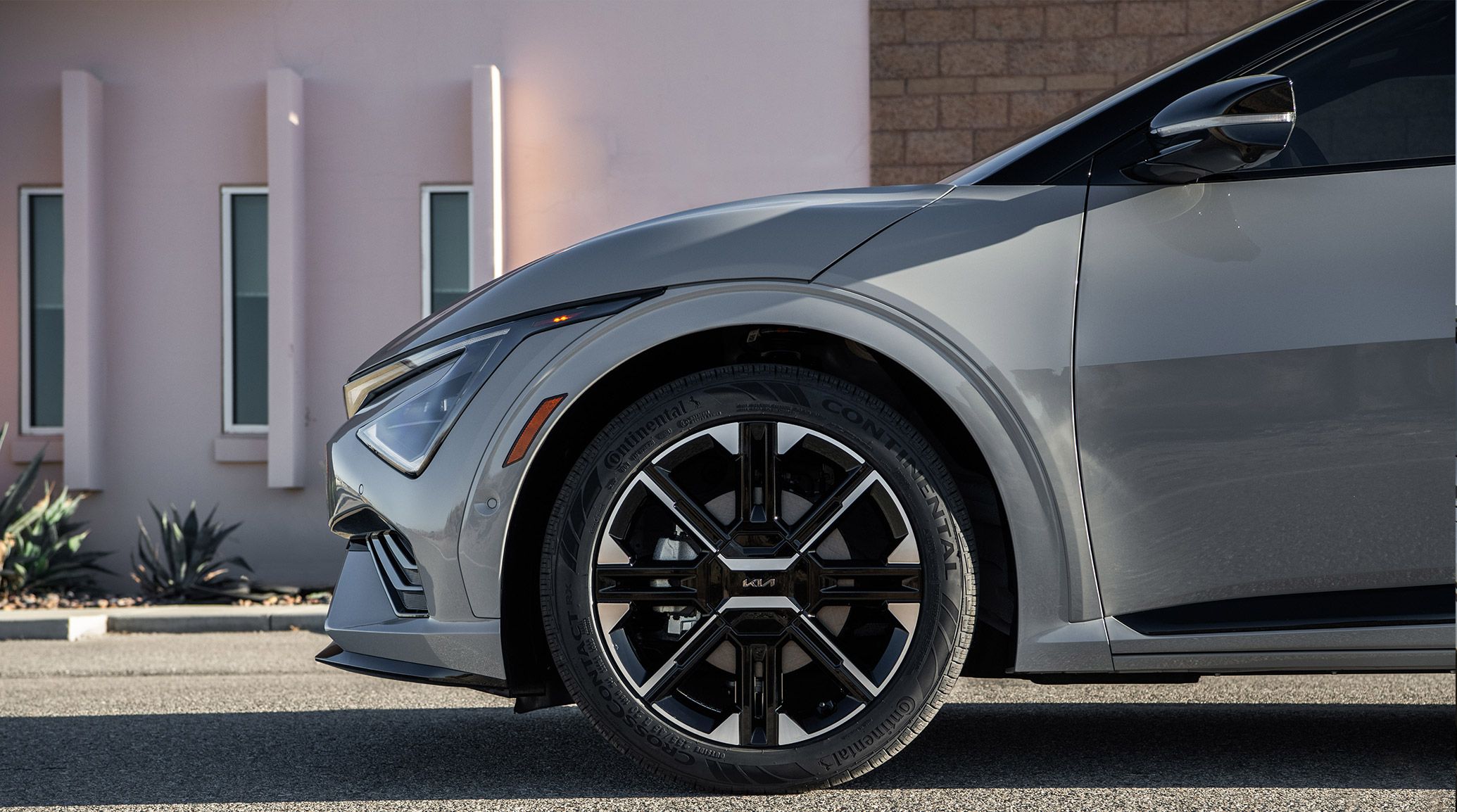 Side view of the 2026 Kia EV6 electric CUV's GT-Line trim with all-wheel drive, focusing on the distinctively patterned 20 inch alloy wheels in black and silver. The vehicle is parked outside of a desert home with brick, windows, and shrubs visible in the background.