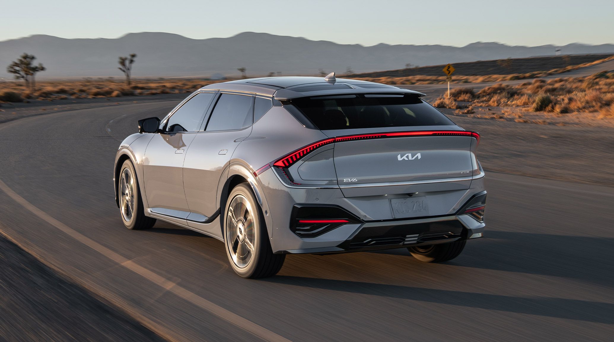 Rear driver-side angle of the 2026 Kia EV6 GT-Line AWD electric CUV in grey, zooming around a curve on a windy desert road, with Joshua trees and mountains visible in the distance. The focus is on the rear fascia and exciting red LED taillights.