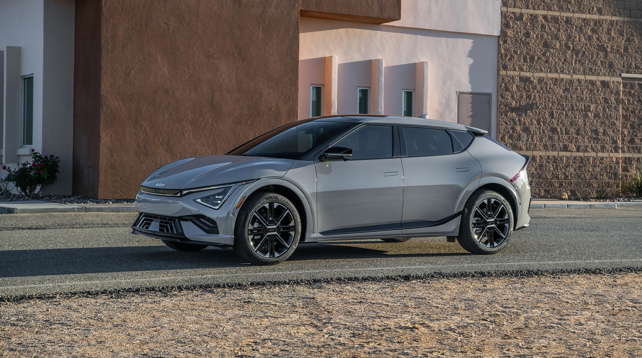 Exterior driver's side photograph of the 2026 Kia EV6 electric CUV in grey, highlighting the sleek shape in the bright sun. The vehicle is parked outside of a desert home on blacktop.