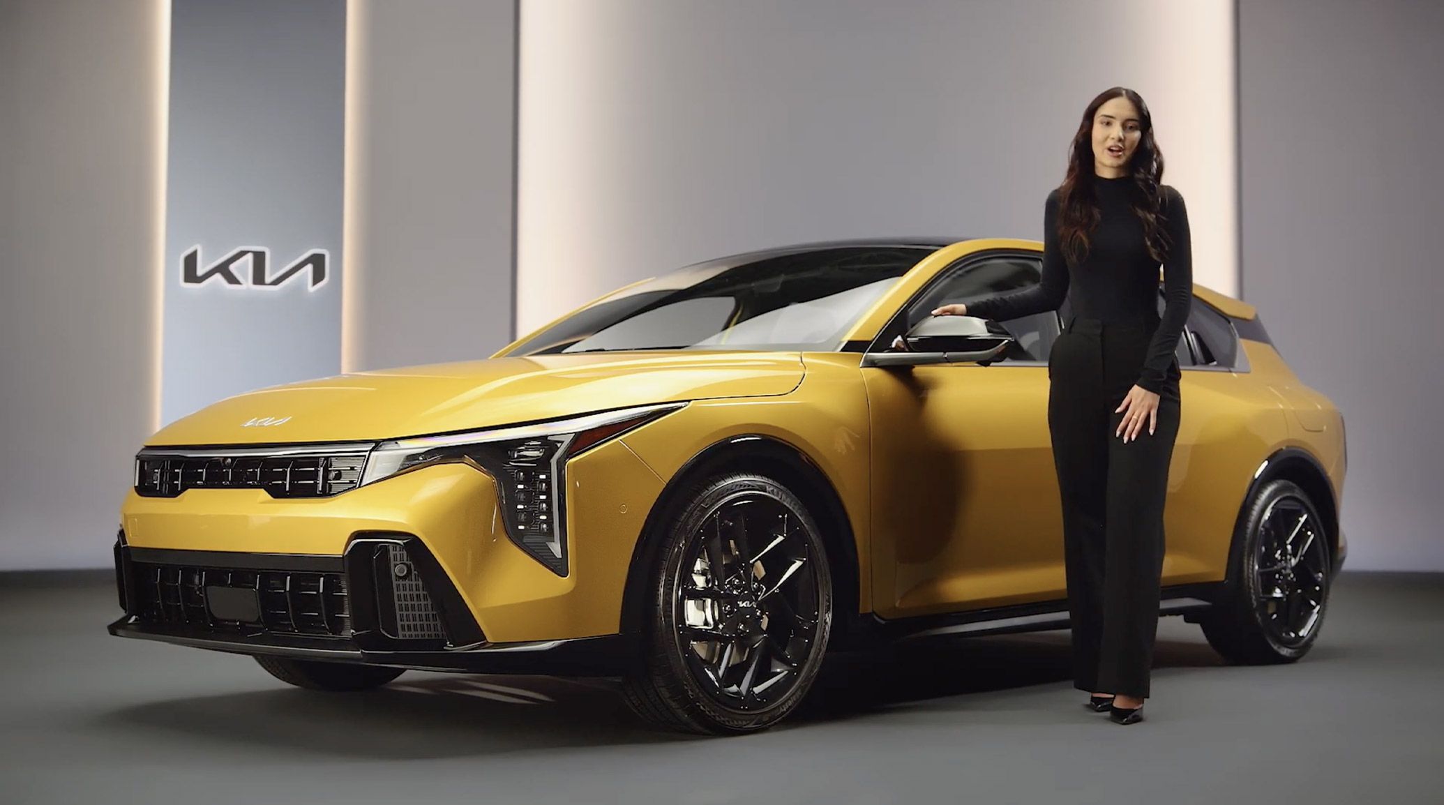 2026 Kia K4 Hatchback in yellow displayed in a white gallery with the Kia Logo on the wall, shown in a front three-quarter view with a brunette female model standing beside the vehicle.