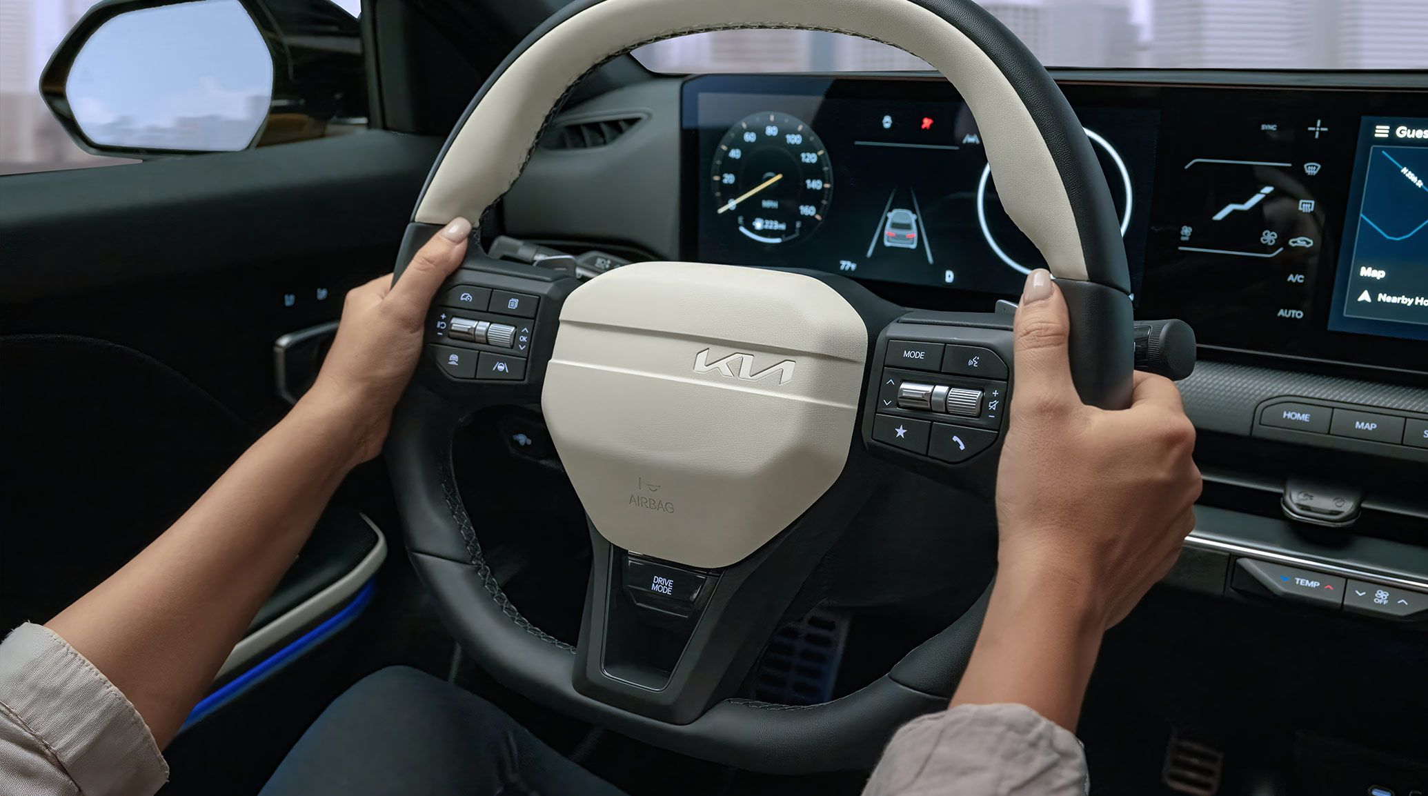 An interior, close up shot of the heated 3-spoke steering wheel on the 2026 K5 Hatchback GT-Line Turbo, showing the driver's hand on the wheel with the digital dashboard that features the speedometer on the left and the vehicle image in a lane in the middle.