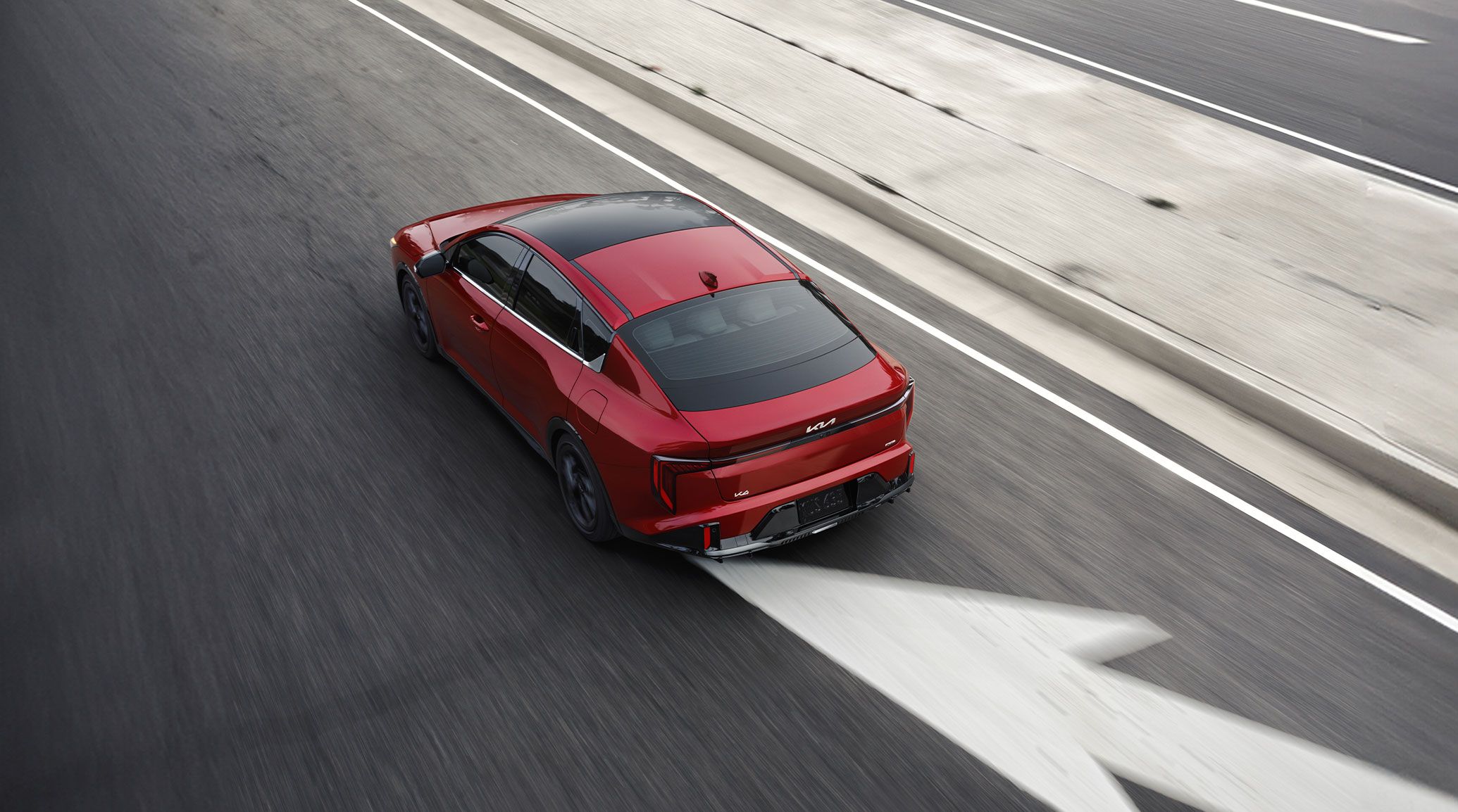Overhead rear view of the 2026 Kia K4 sedan in red, driving down a concrete highway in a merging traffic lane