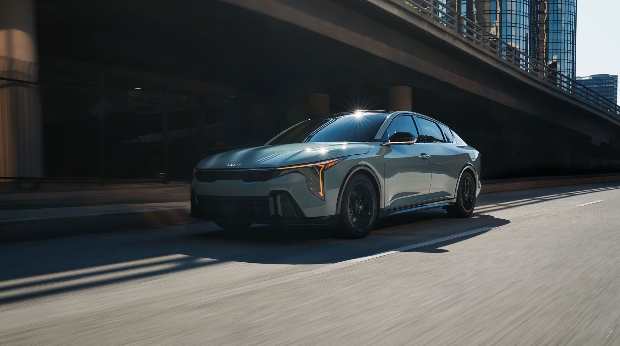 Driver-side front three-quarter view of the 2026 Kia K4 sedan GT-Line trim driving in an urban environment, featuring its unique front bumper with gloss black trim
