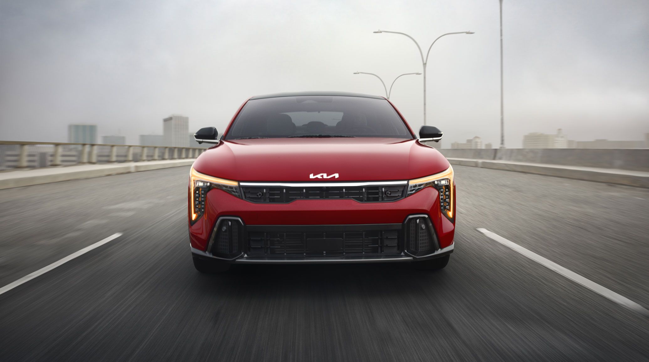 Front view of the 2026 Kia K4 sedan driving straight towards the camera, showing its windshield, hood, front grille, and amber daytime running lights with cube projector headlights on the GT-Line Turbo trim in red.