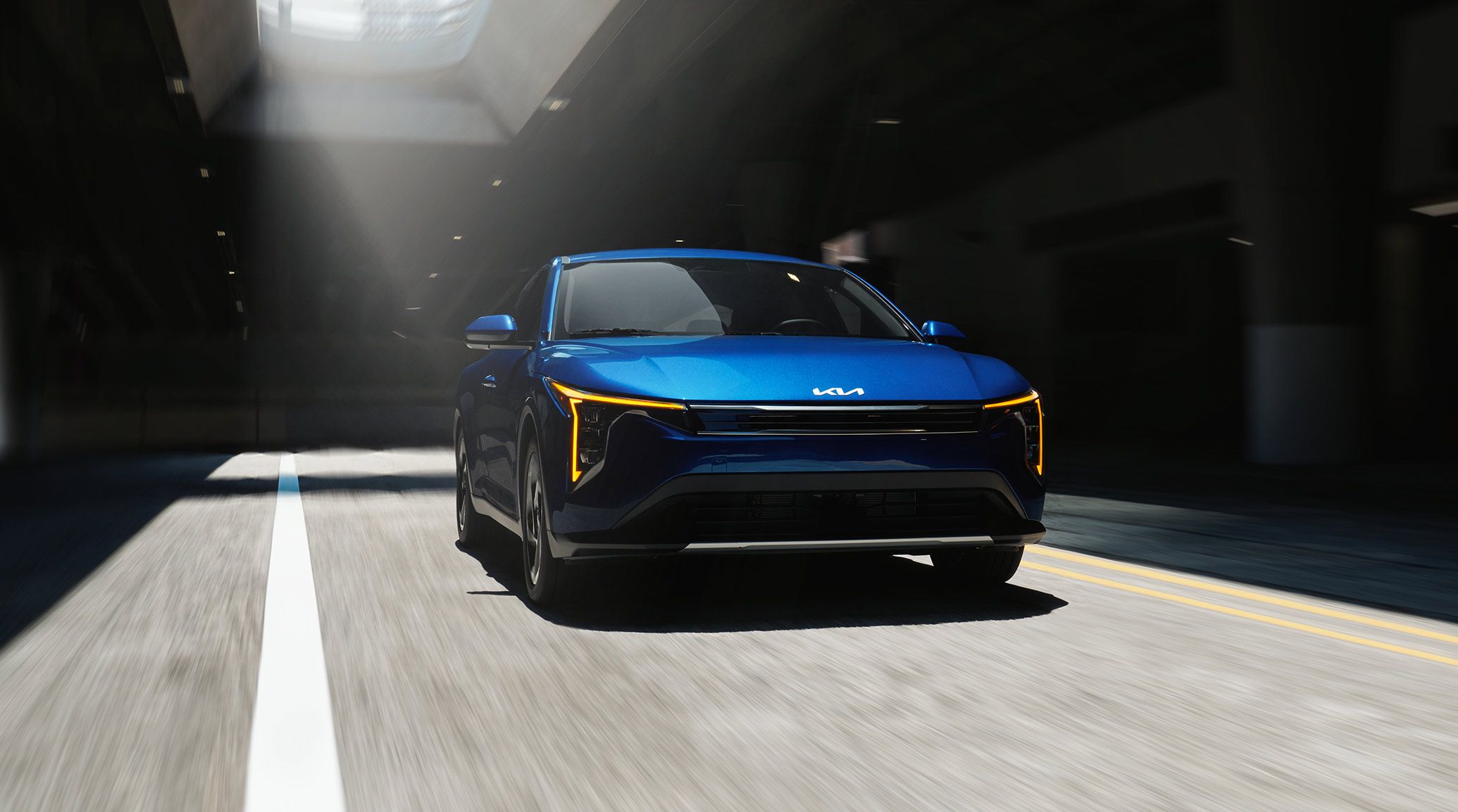 Front view of the 2026 Kia K4 sedan EX trim in vibrant blue with dynamic light and shade effects, featuring the gloss black and chrome trim grille, as it drives through an underground concrete structure