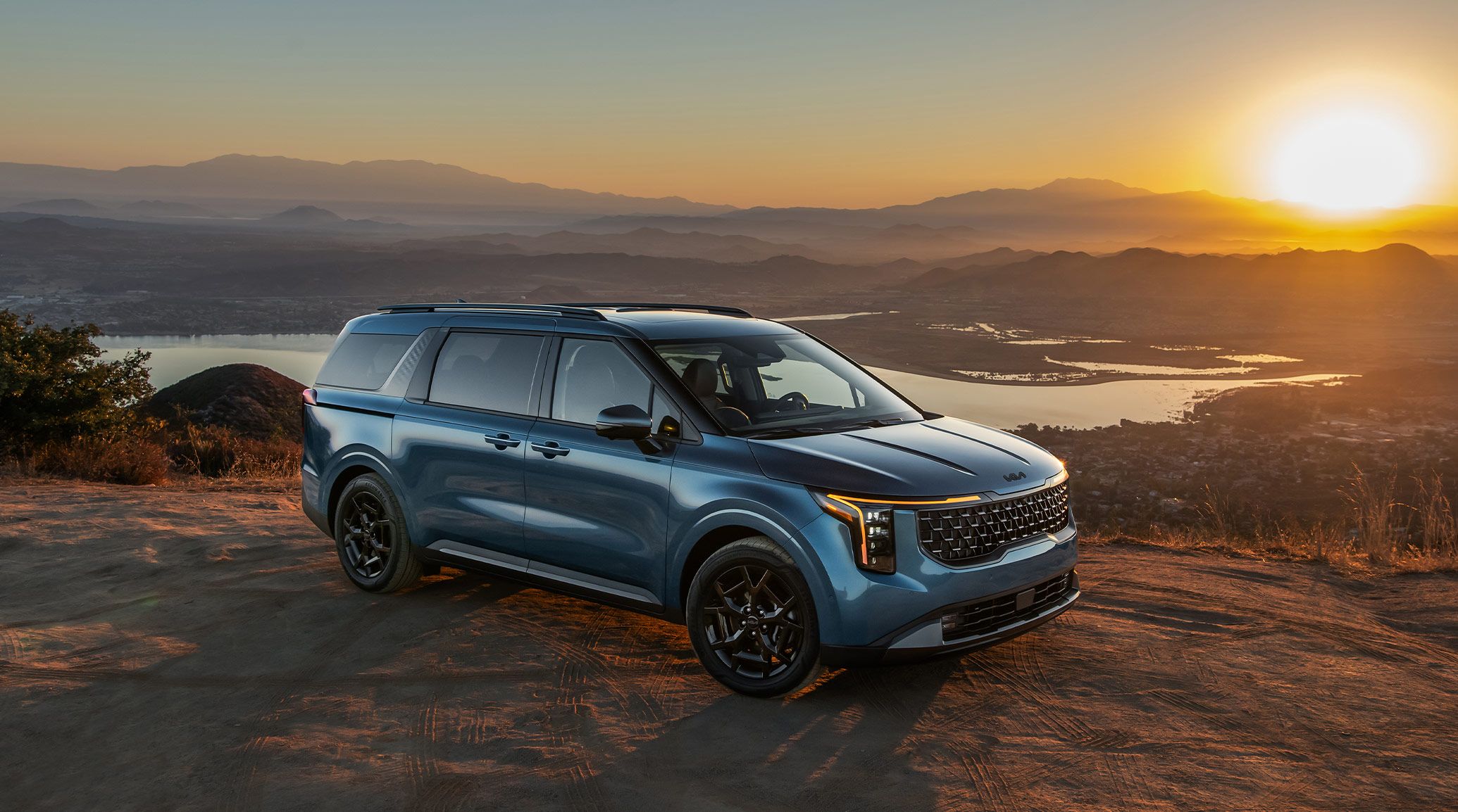 A front three-quarters view of the 2027 Kia Carnival Minivan in blue parked on a dirt road with a lake and mountains visible behind, highlighting its roof rails and bold front grille.