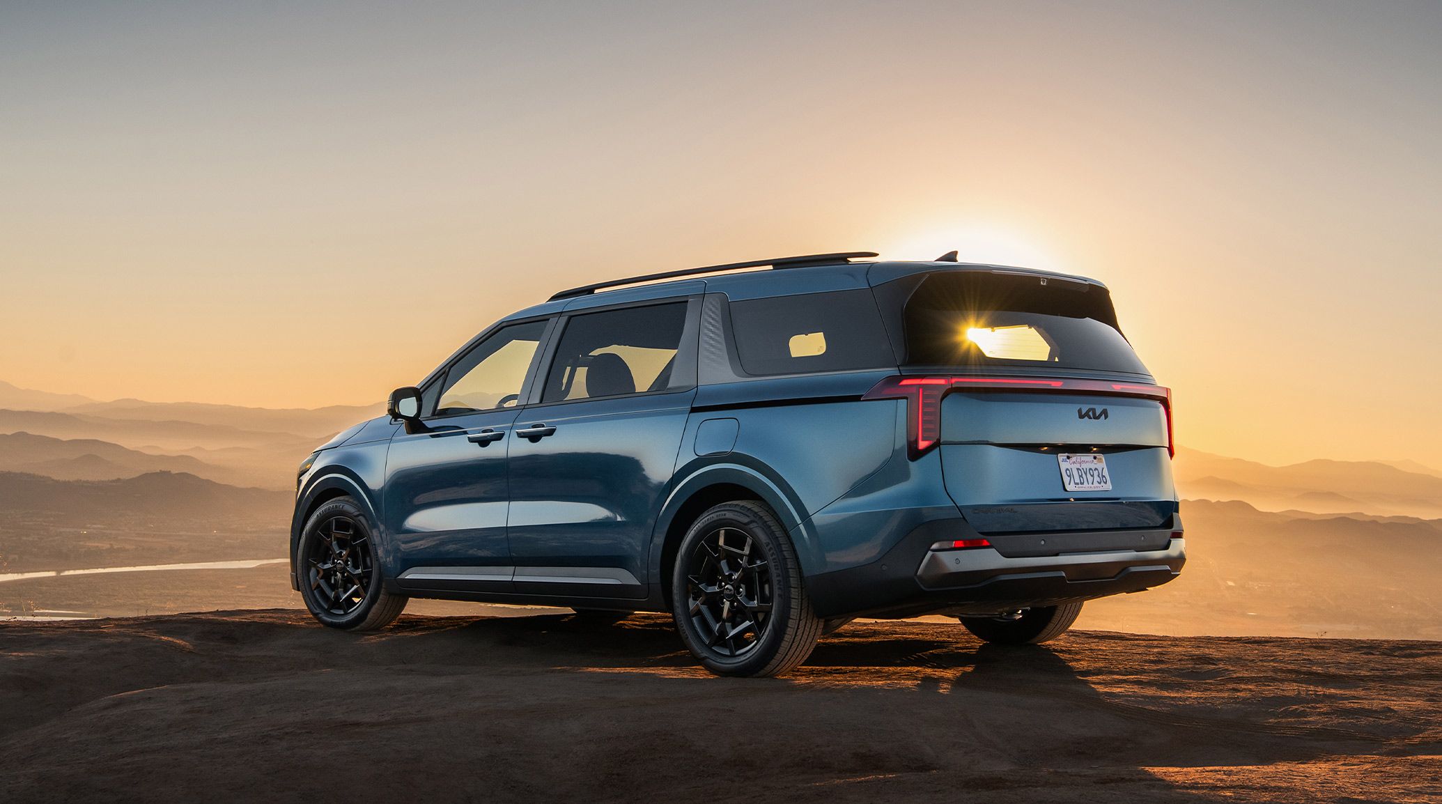 The 2027 Kia Carnival MPV in blue is shown from the rear, parked outdoors on a cliff against a sunset with mountains behind. The image highlights its LED taillights and purpose-built roof rails.