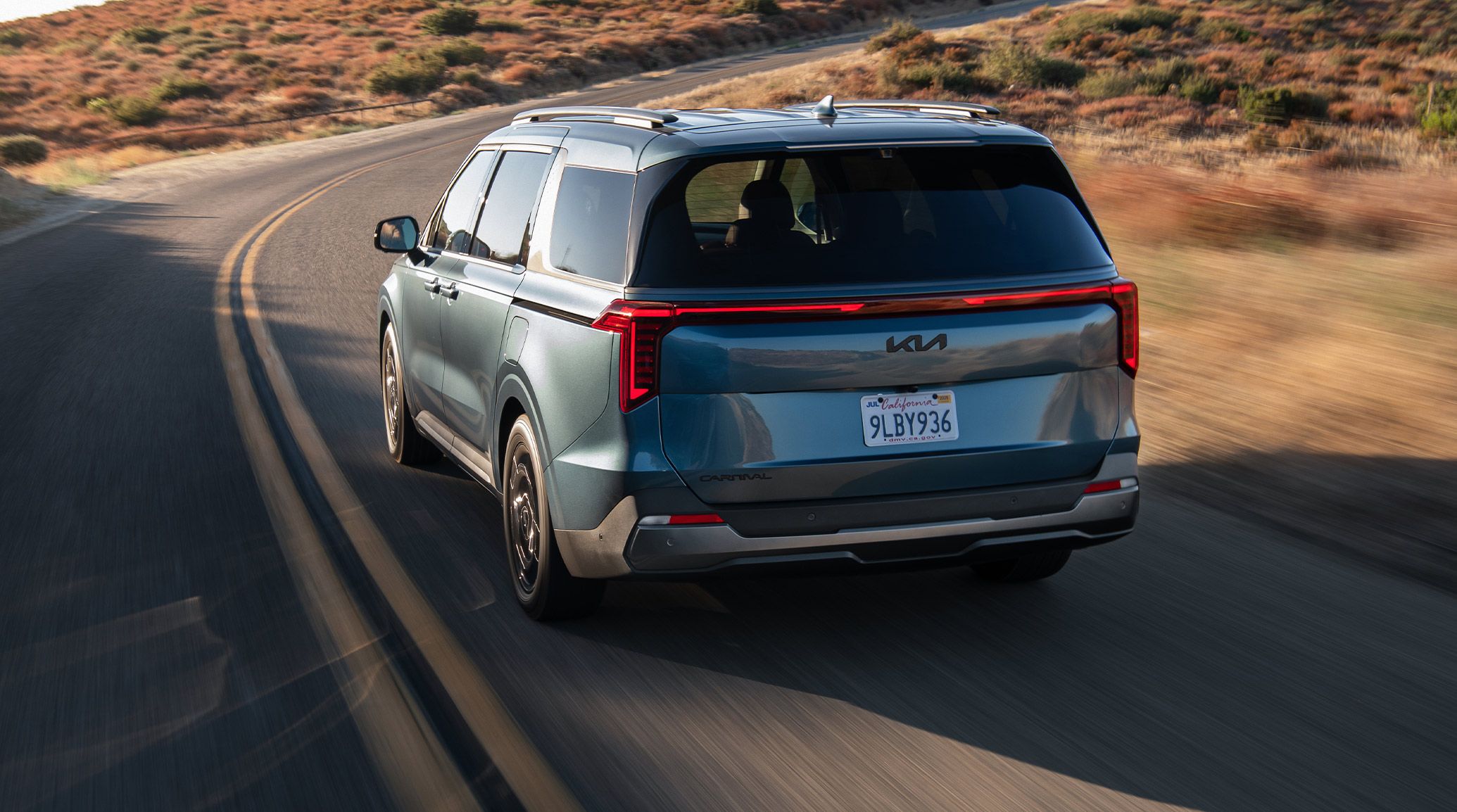 A rear image of the 2027 Kia Carnival, SUV-inspired minivan, driving along a winding road with bush blurred in the background, highlighting its full-width LED taillights and modern design.