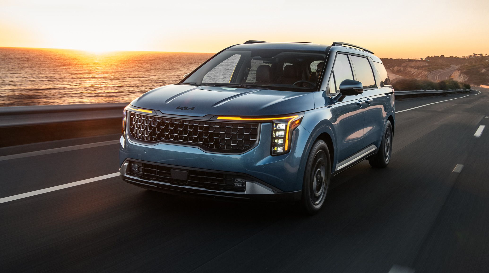 2027 Kia Carnival MPV SX Prestige in blue, three-quarter front view, highlighting the vertical LED headlights, sleek grille, and mirrors. The SUV-inspired minivan is driving along an oceanfront highway during sunset.