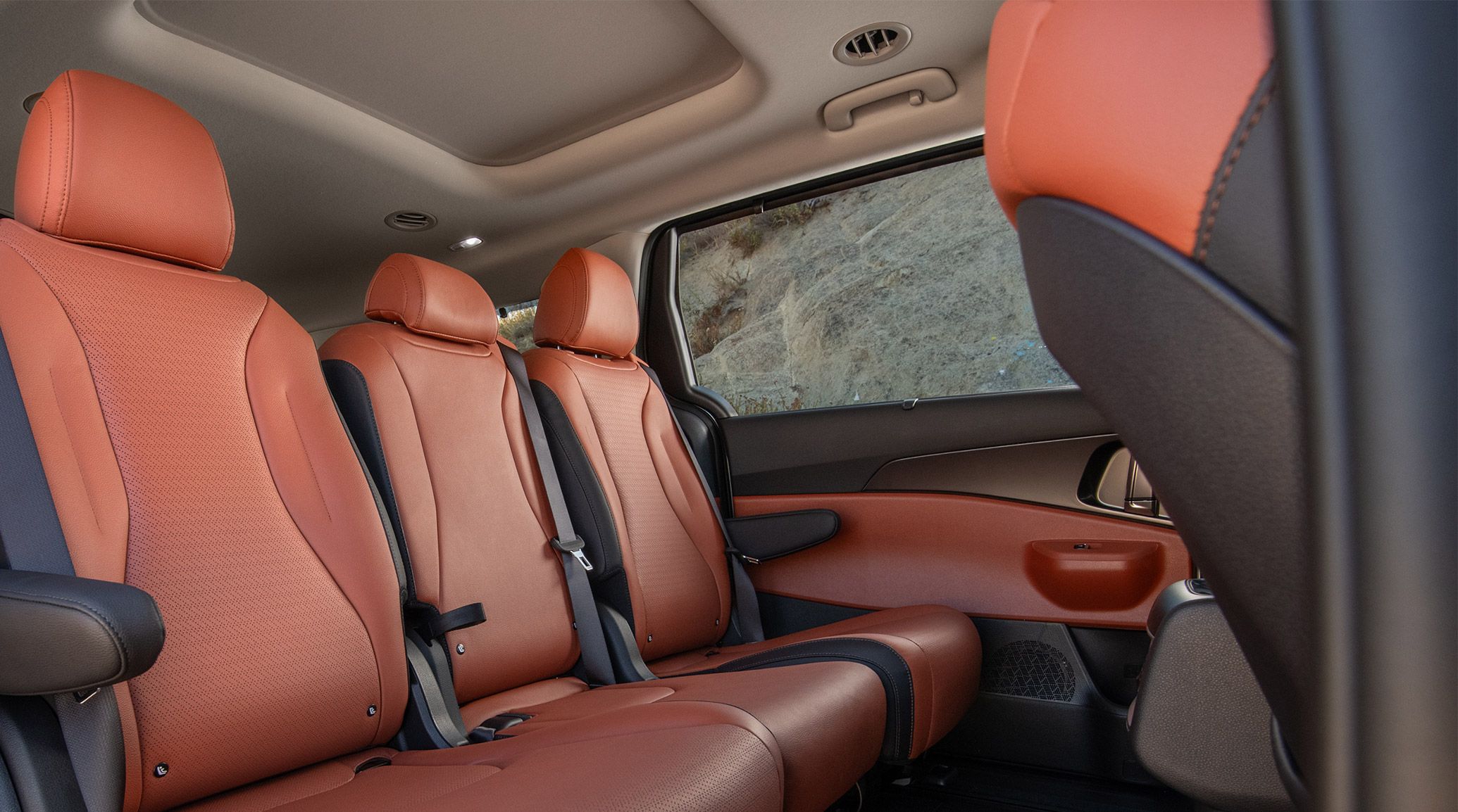2025 Kia Carnival MPV interior view of the spacious 2nd-row seats, featuring a two-tone red and black leather seat trim, with shrubs on a hill peeking through the window.