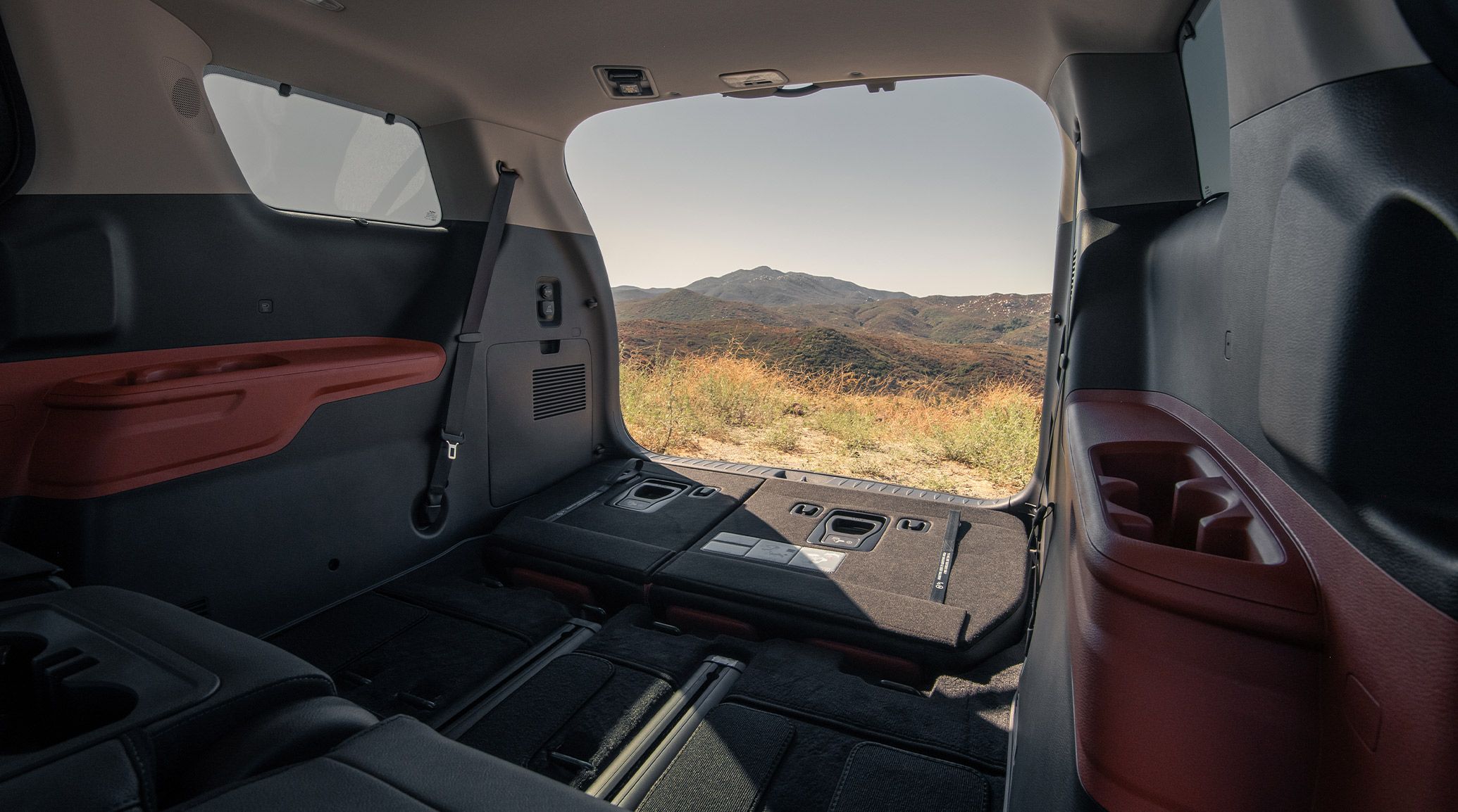 Spacious trunk view of the 2027 Kia Carnival MPV with foldable seating placed down and rear hatch opened overlooking a scenic landscape of plants and mountains.