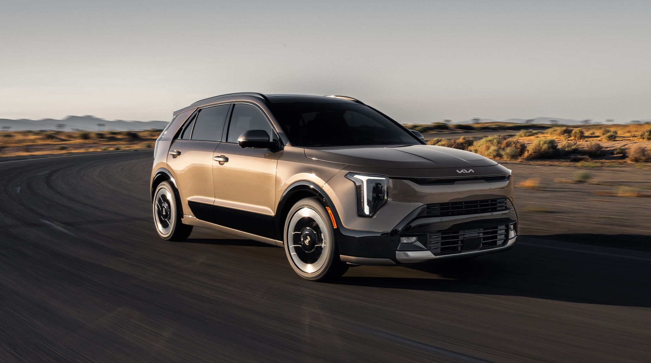 Front passenger side angle photograph of the 2027 Kia Niro Hybrid Electric SUV driving around a curved desert road in the sun.