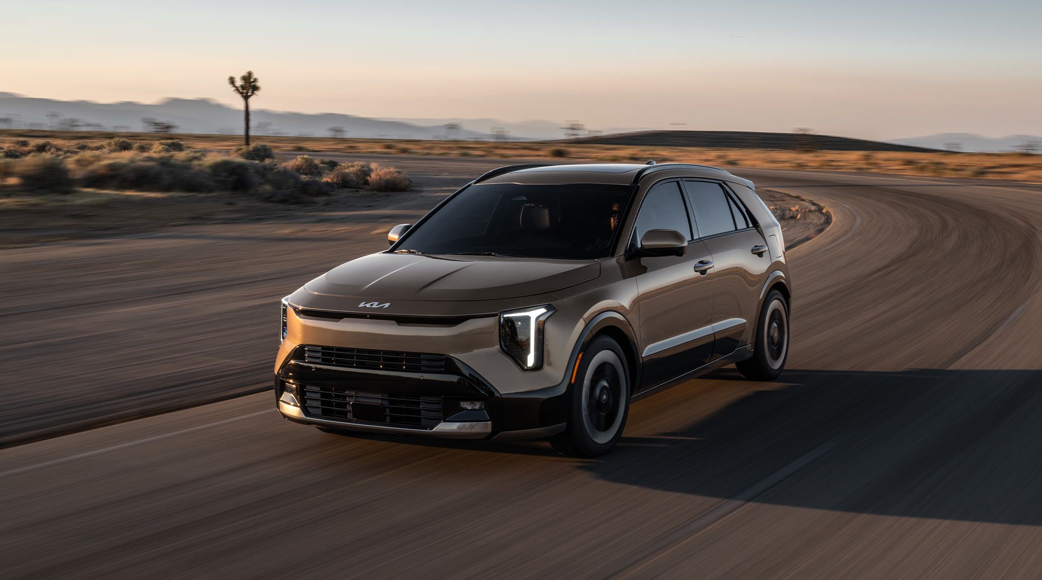 Dynamic front driver's-side angle view of the 2027 Kia Niro Hybrid subcompact SUV in bronze brown driving quickly around a curve in a road in the desert. It looks impressive and powerful.