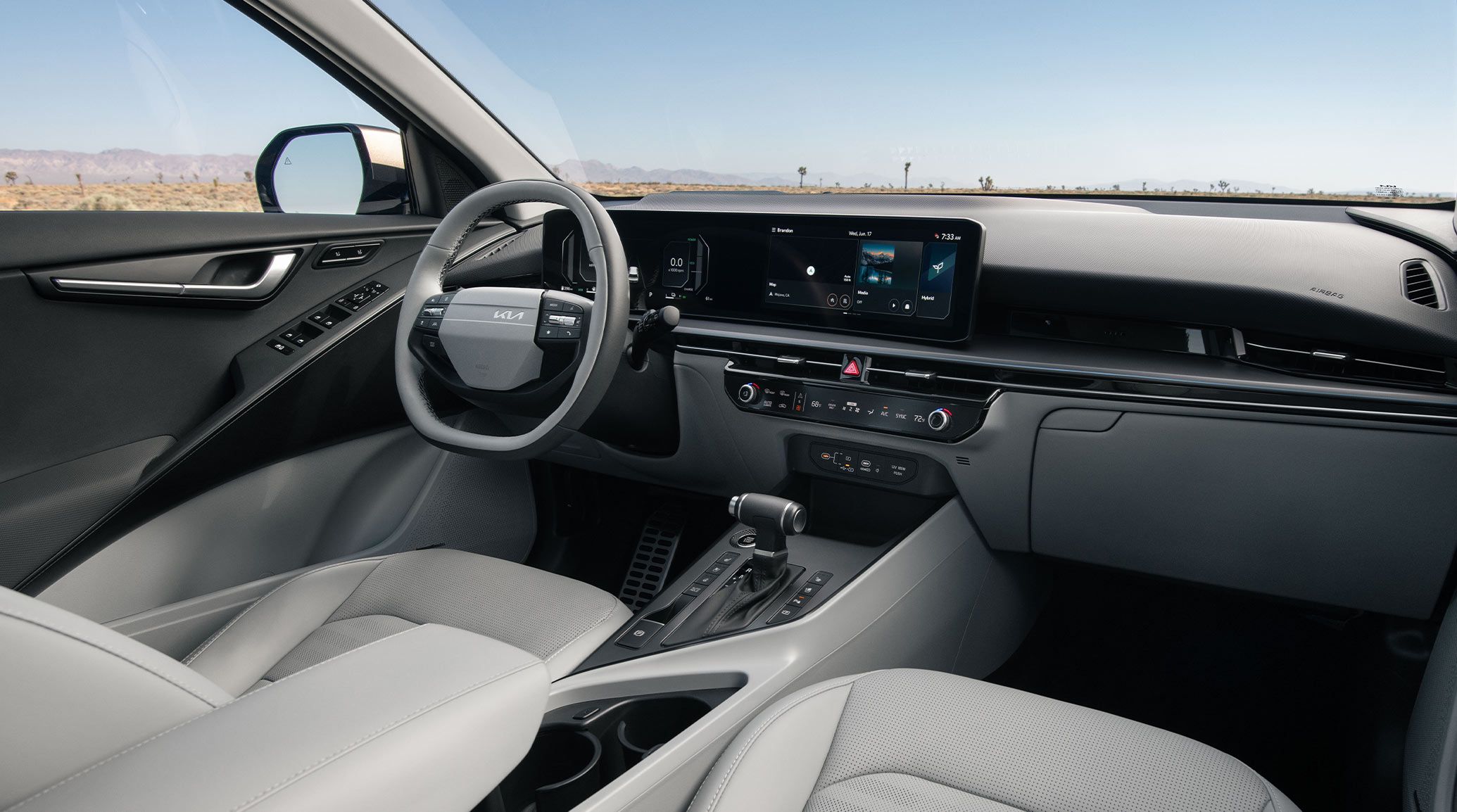 Interior view of the 2027 Kia Niro Hybrid SUV showcasing the steering wheel, grey front seats, center console, and dashboard. Desert landscape is visible through the windshield.