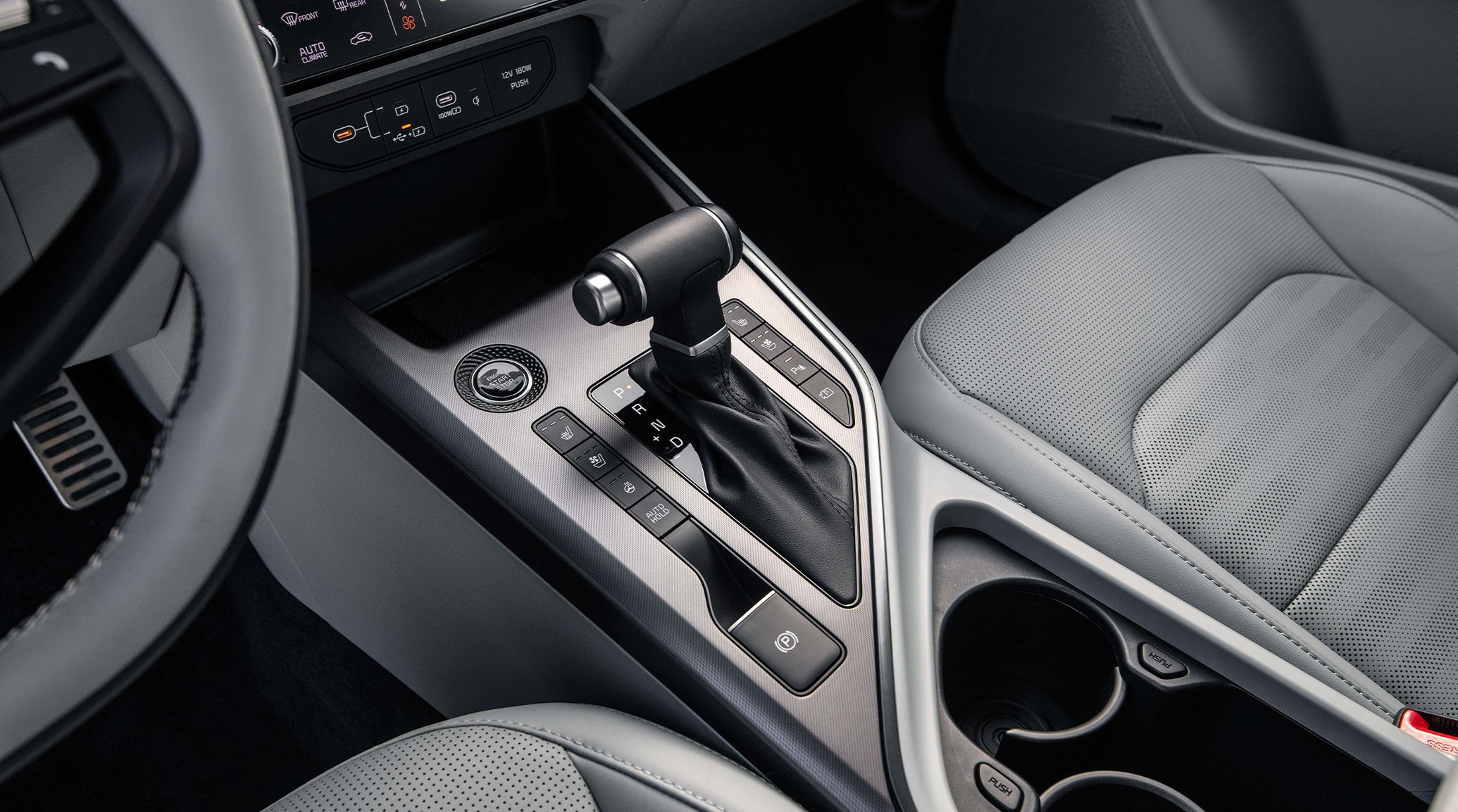 Interior view of a Kia vehicle showing the shifting stick in the center console, as well as cupholders, buttons, and front seat fabric details.