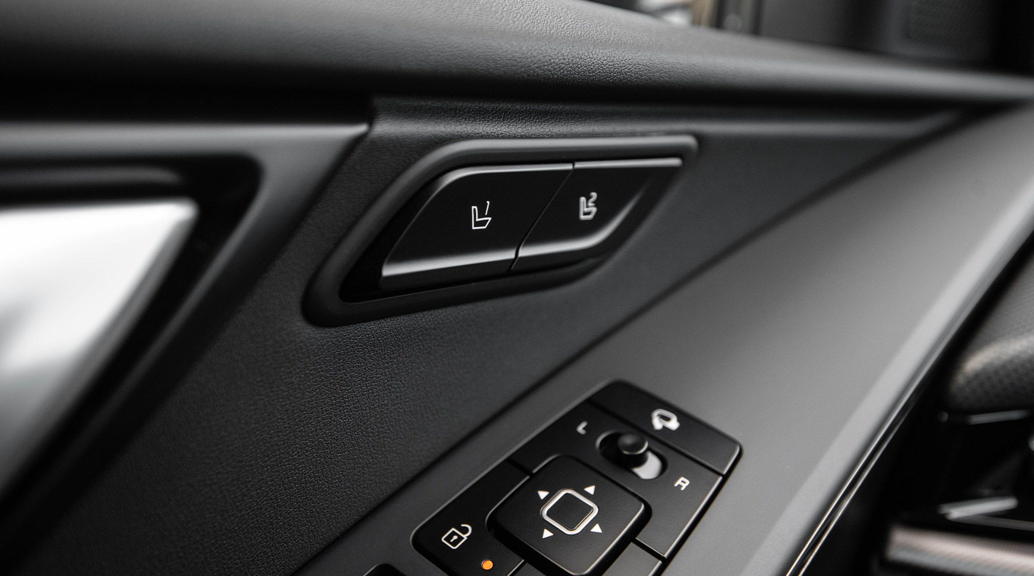 Close up photograph inside the 2027 Kia Niro Hybrid Electric SUV showing buttons on the driver's door, including arrow buttons, a lock button, a left-right switch, and most notably, two buttons with numbered seat memory icons.