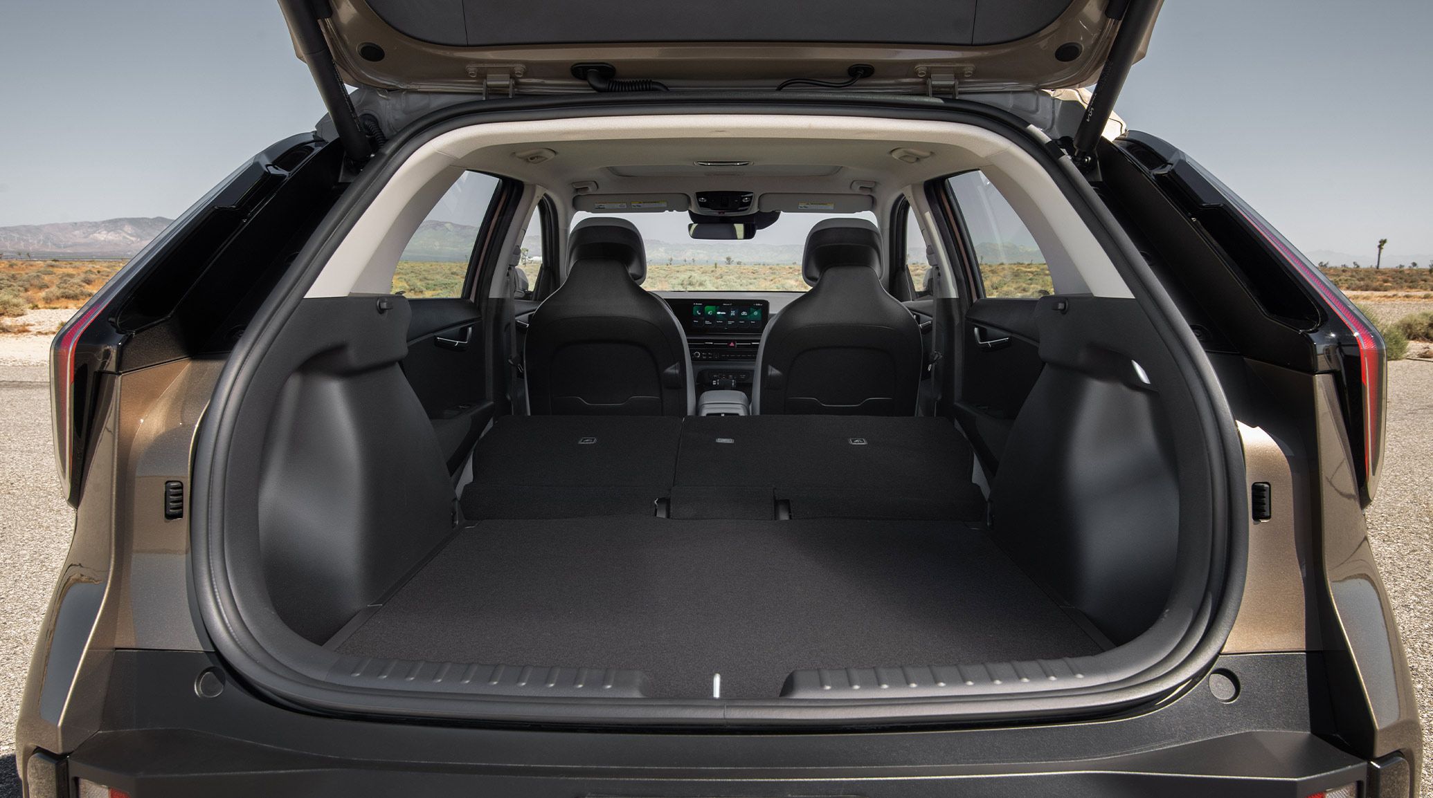 The 2027 Kia Niro Hybrid Electric SUV with its trunk door opened up, showing the ample empty cargo space available with the second row of seats in the downward flat position. A desert is visible outside the vehicle in the background.