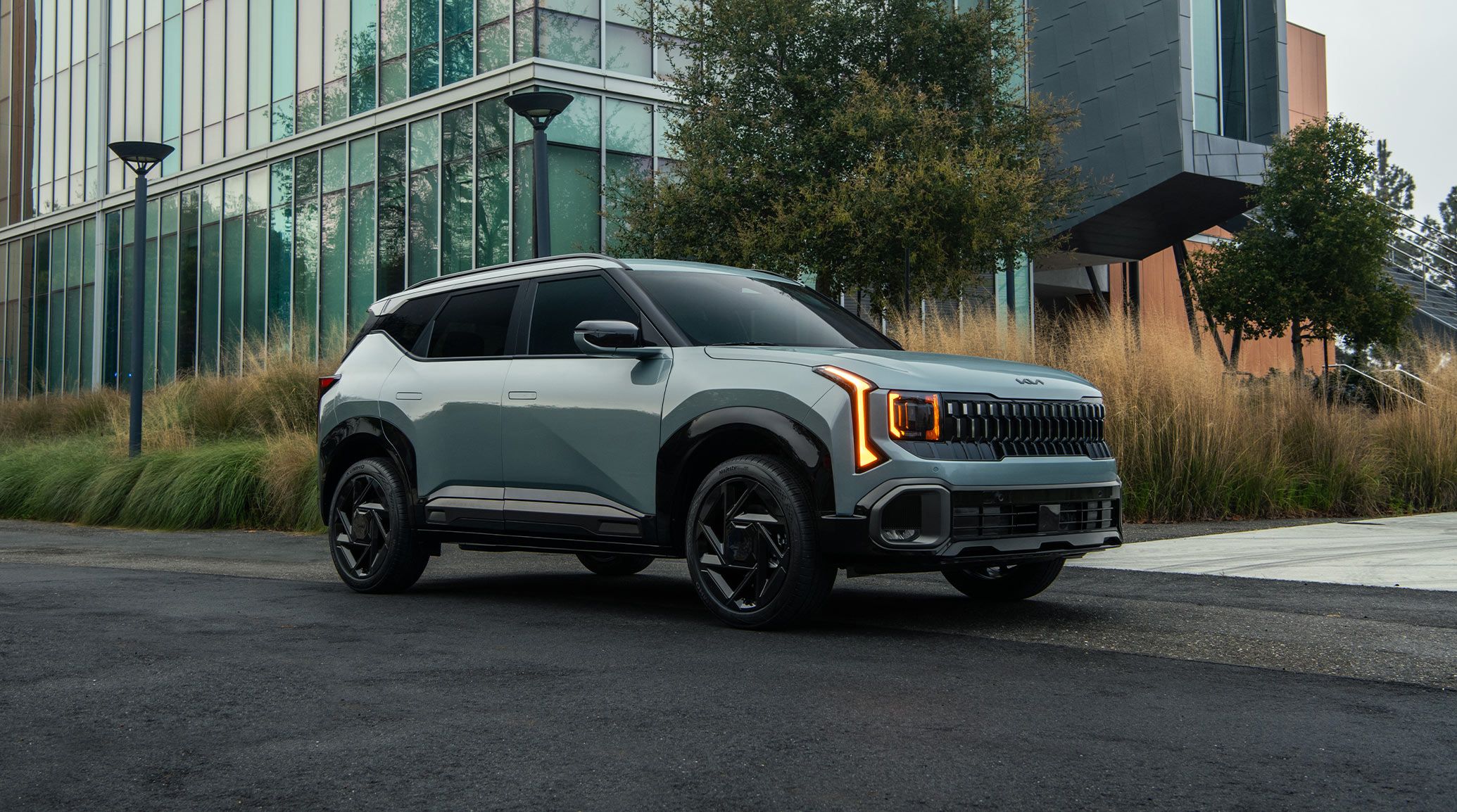 Side view of a blue 2027 Kia Seltos Hybrid compact SUV parked on a road with pants and modern buildings behind the vehicle. This photograph shows the gloss black exterior trim, as well as the bright orange LED headlights.