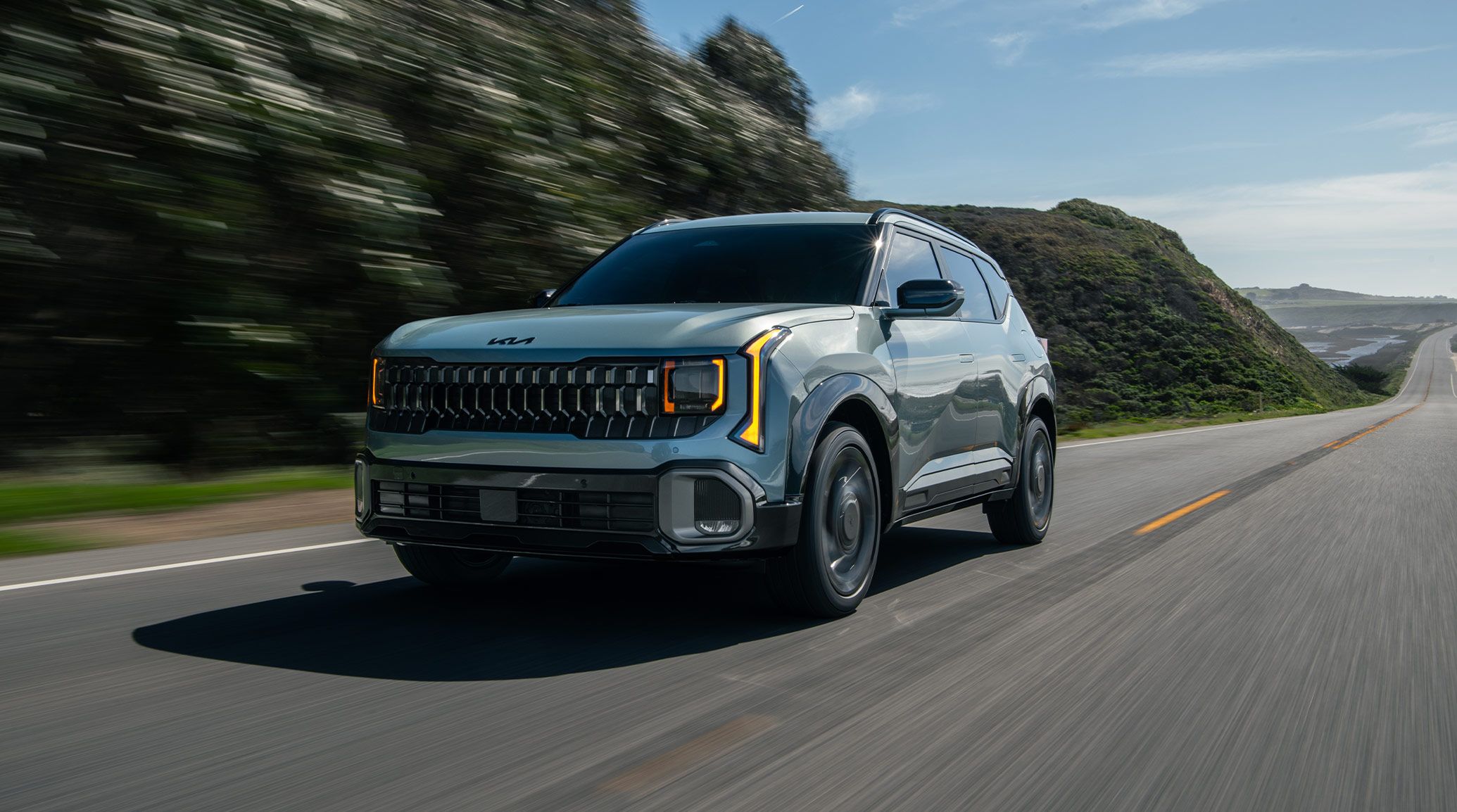 Shot of the 2027 Kia Seltos Hybrid in blue driving quickly through a road surrounded by green hills. The image highlights the compact SUV's bold front grille, tinted windshield, and LED headlights.