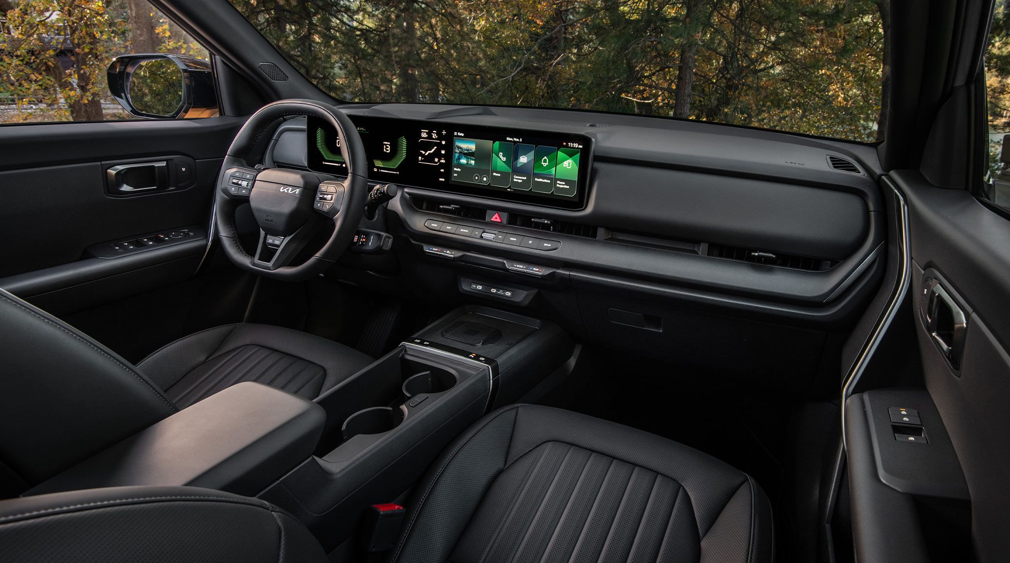 Interior shot of the 2027 Kia Seltos Hybrid in black from the passenger seats, showcasing its steering wheel, dashboard display, center console, glove compartment, and more. Trees are visible through the windshield.