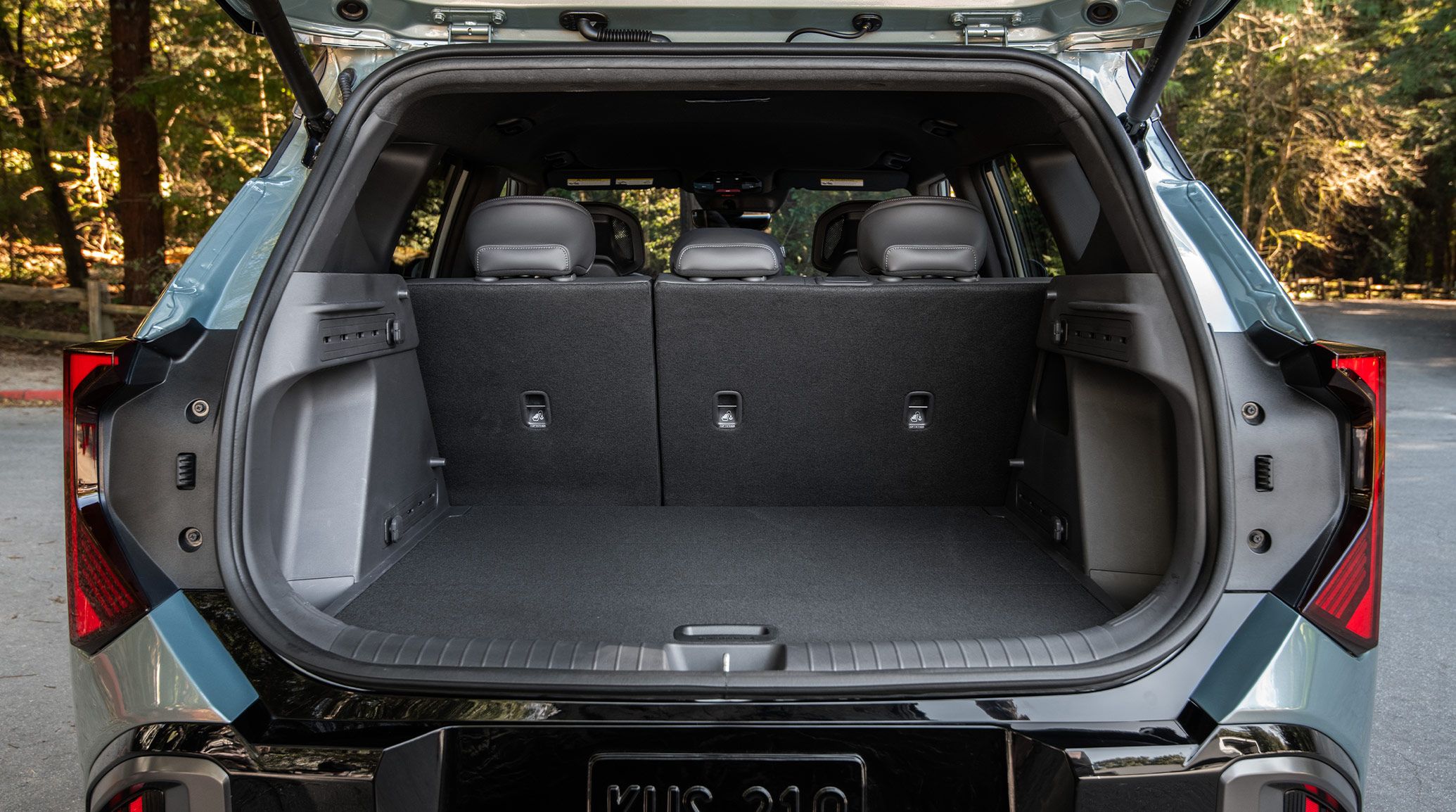 A rear photograph of the 2027 Kia Seltos Hybrid showing the trunk open and with the 2nd-row seats folded up, highlighting the roomy cargo space the vehicle offers. The compact SUV is parked on the road with trees in the background.
