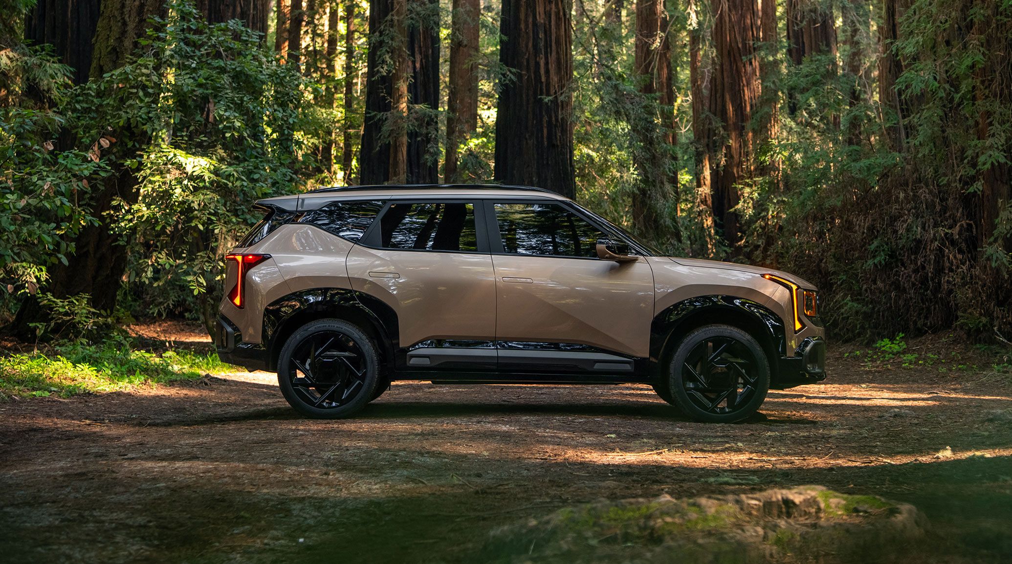 2027 Kia Seltos X-Line SX AWD in brown parked in a beautiful forest setting, shown in full side profile with black wheels and rugged styling highlighted.