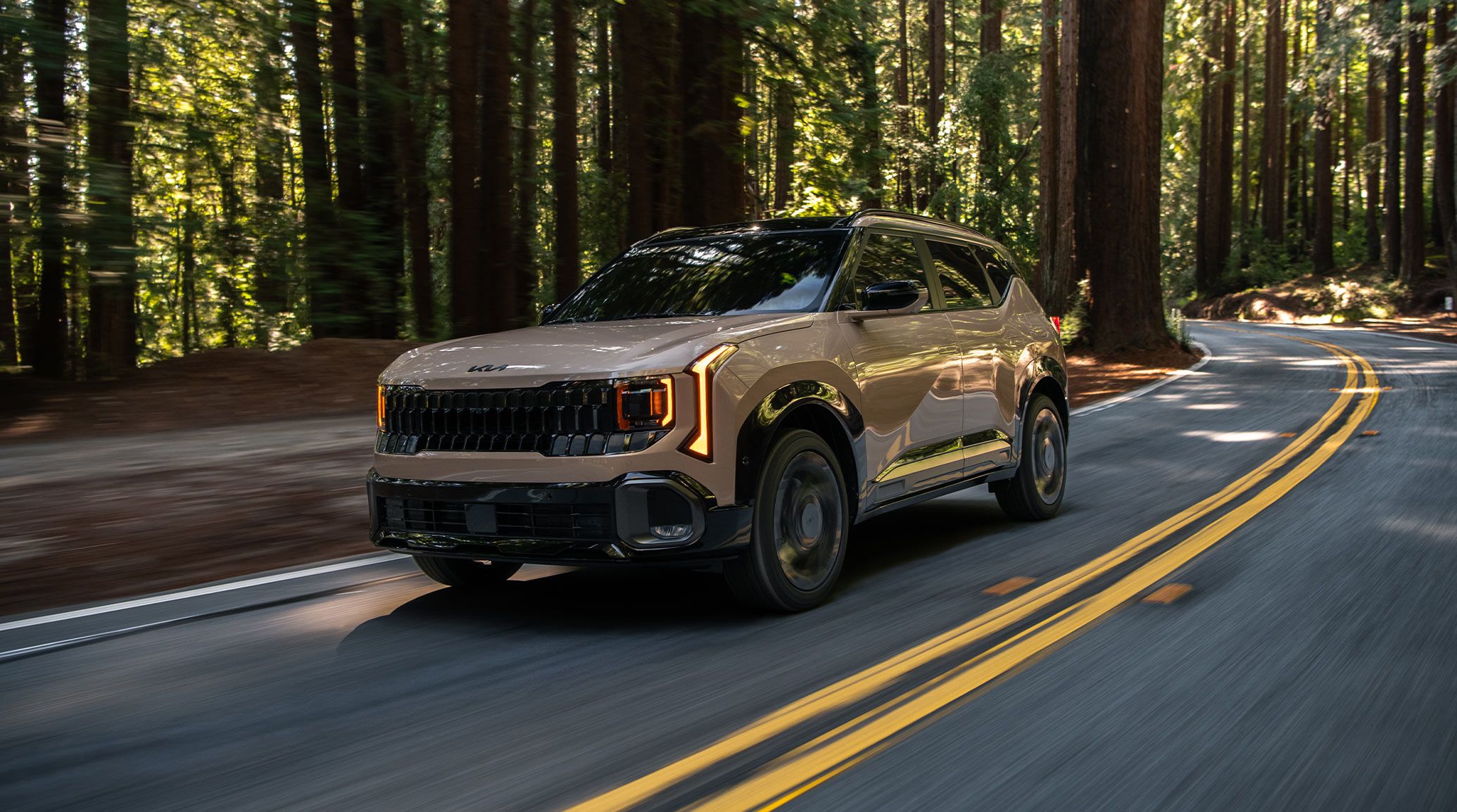 Shot of the 2027 Kia Seltos in brown driving quickly through a winding forest road. The image is highlighting the compact SUVs bold front grille, tinted windshield, and LED headlights.