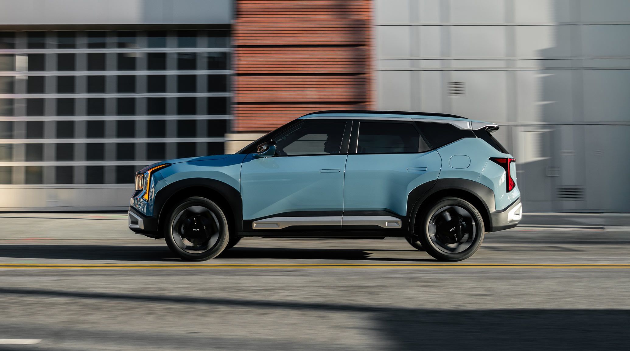 The 2027 Kia Seltos in blue driving through an urban setting, shown in full side profile with black wheels and chrome accent trims highlighting its modern exterior design.
