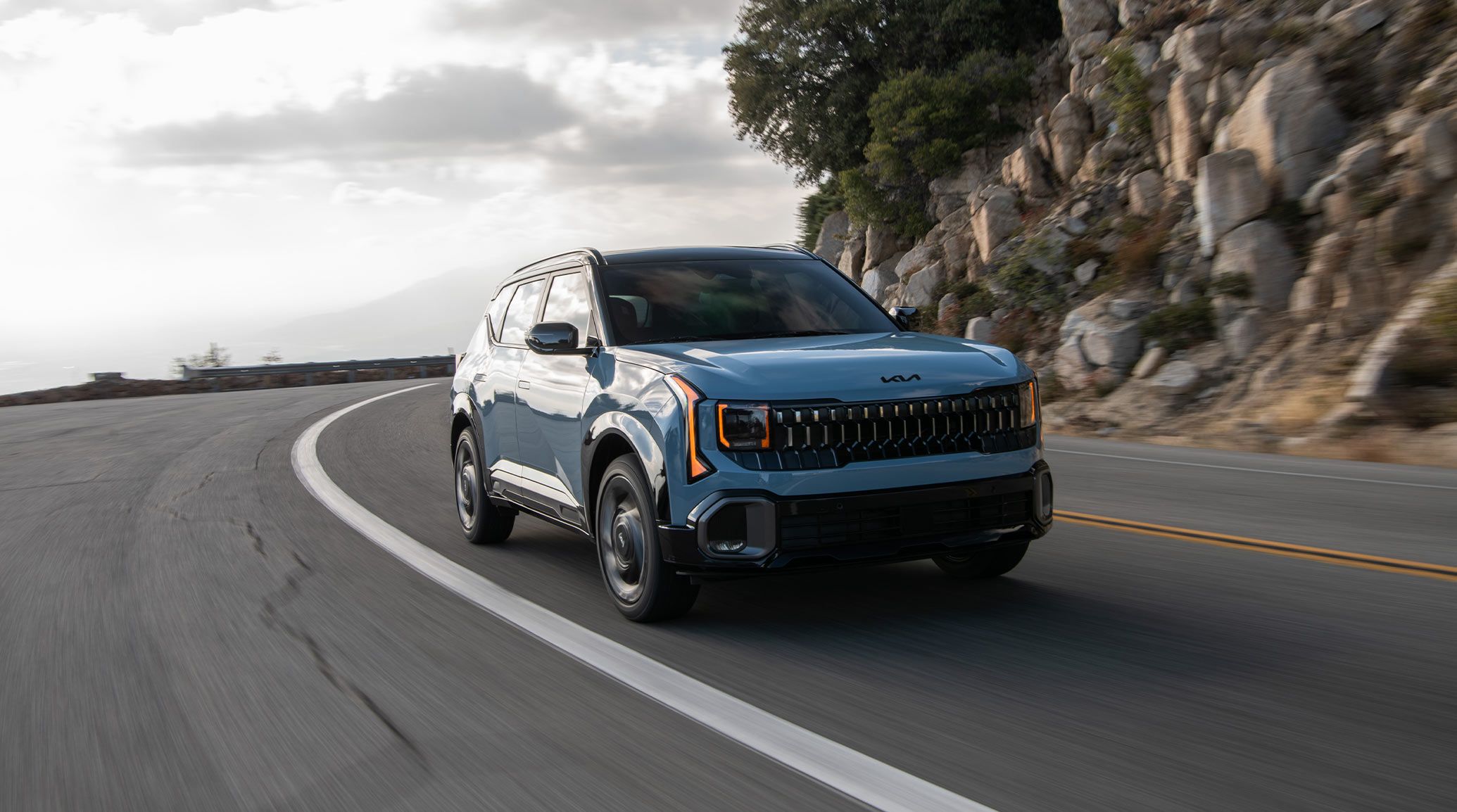 The 2027 Kia Seltos in blue driving along a winding mountain road with cloudy skies, highlighting its unique front fascia and black grille.