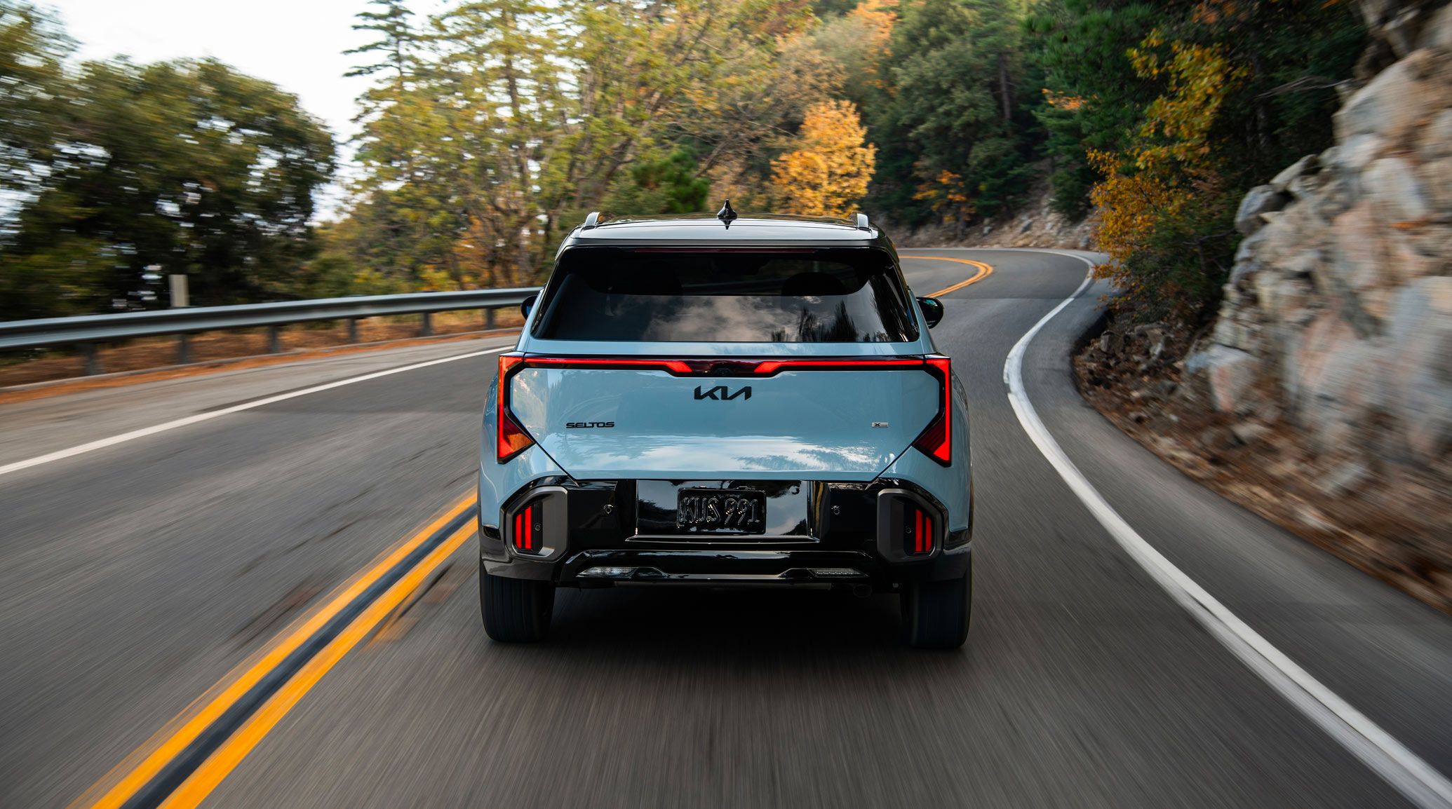 Rear shot of the 2027 Kia Seltos in blue driving rapidly over a road with trees and rocks in the background, highlighting the LED taillights and black emblems.