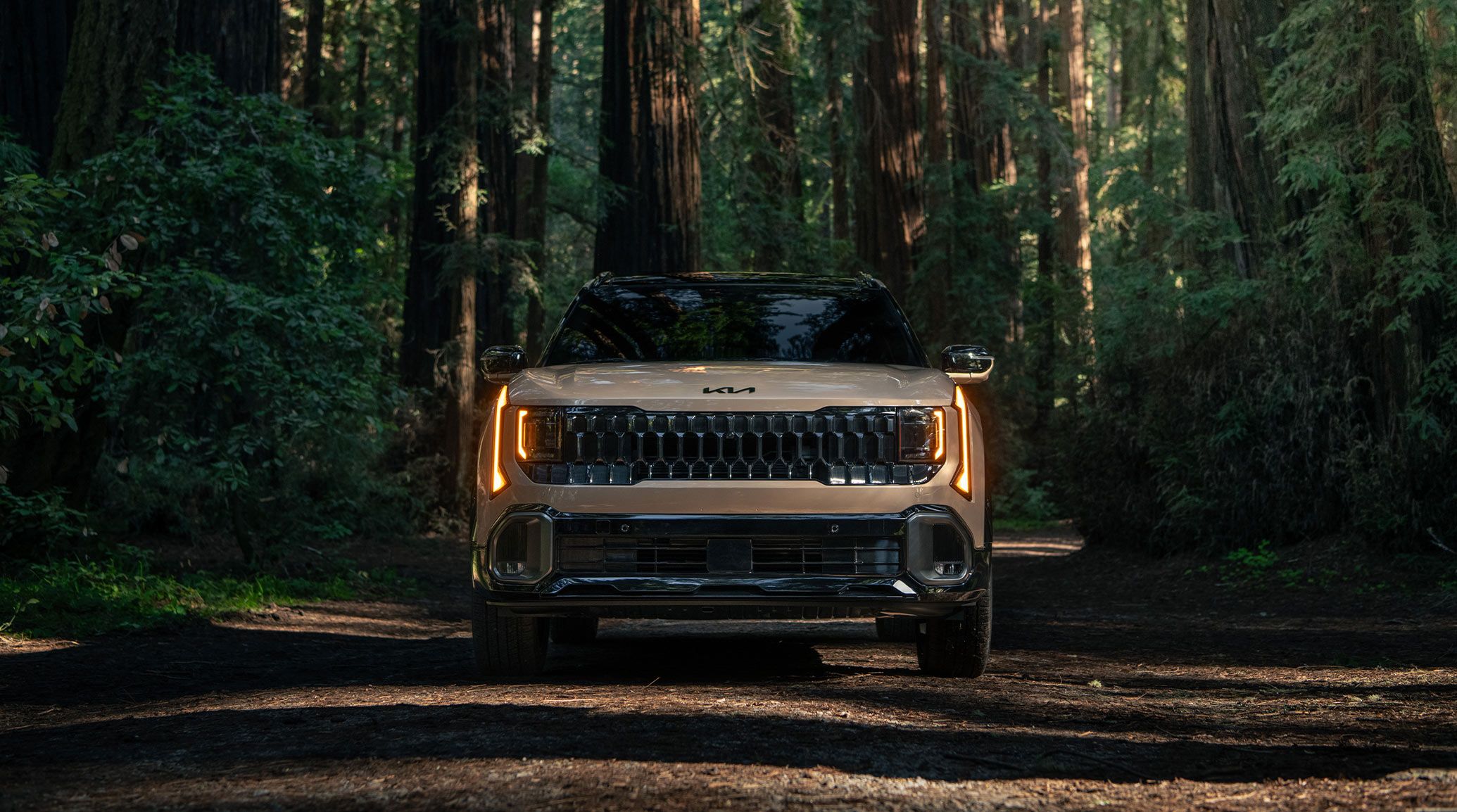 Front view of the 2027 Kia Seltos in brown, with bright LED headlights, showcasing its distinctive grille and front features, including the Kia logo in black. It's parked in an expansive forest.