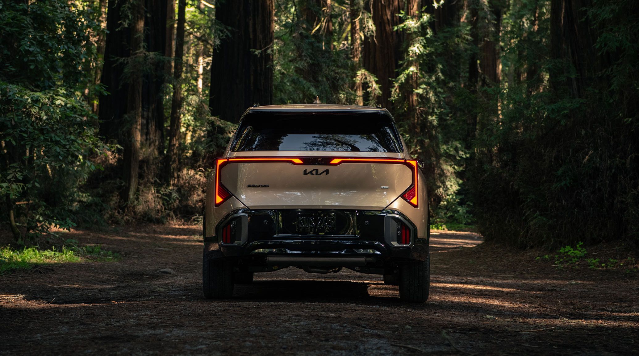 Rear shot of the 2027 Kia Seltos compact SUV in brown, showing off its LED taillights and gloss black trim. It's parked in a dense forest full of very large trees.