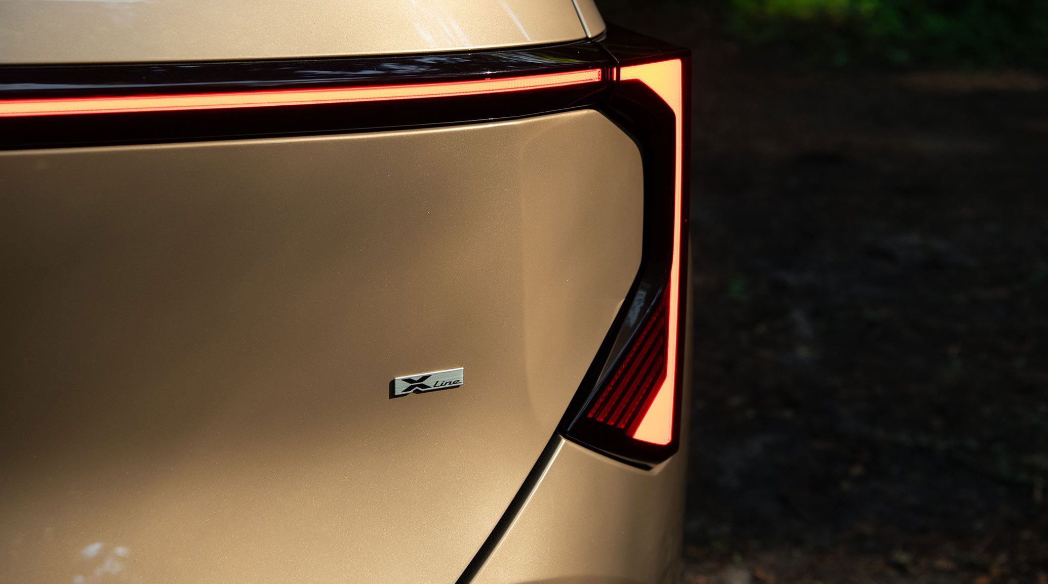 A close-up shot of the 2027 Kia Seltos in brown, highlighting the LED taillights and the silver X-line badge shown underneath the lights. The compact SUV is parked with blurred dirt visible in the background.