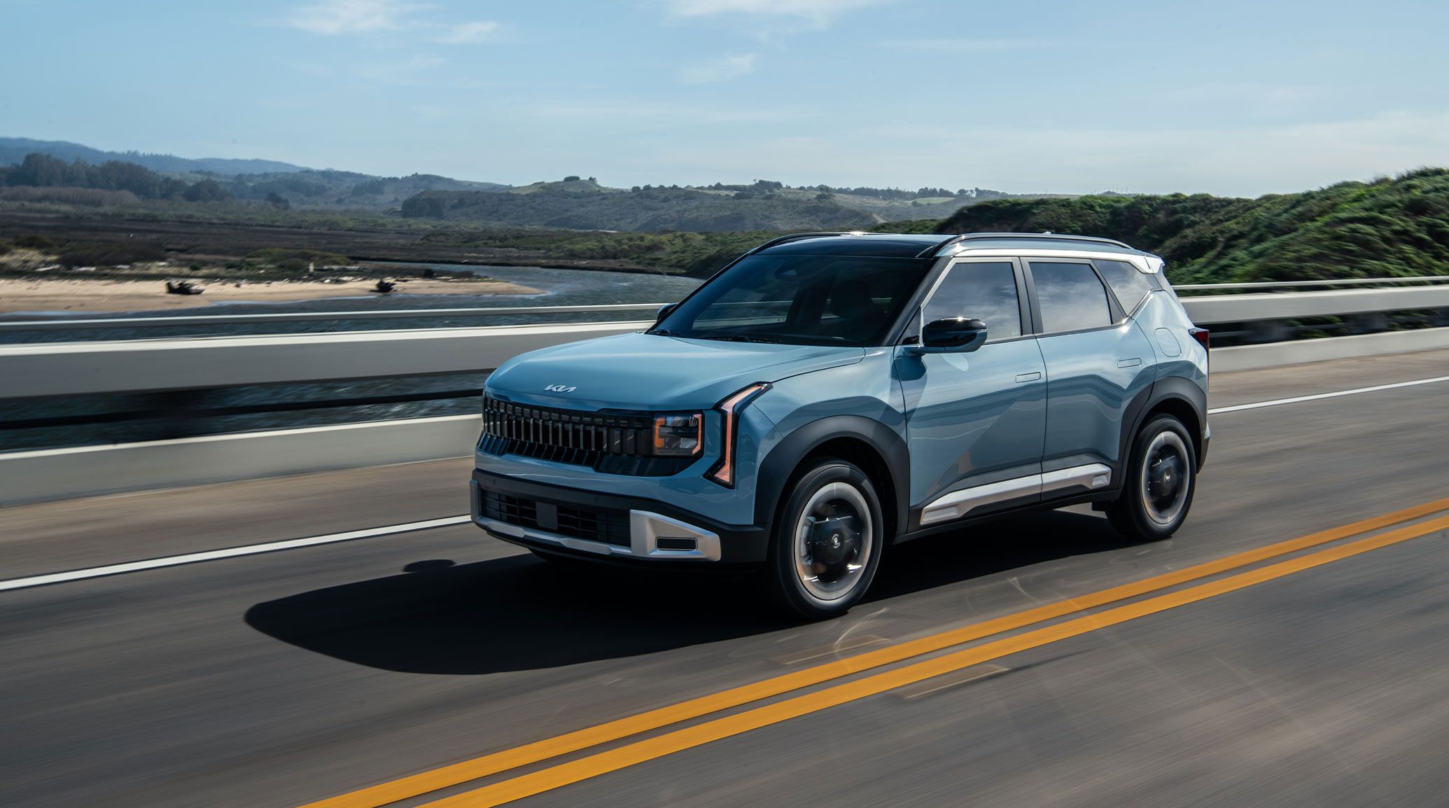 2027 Kia Seltos EX in blue driving on an open highway, shown in front three-quarter view with sleek exterior styling, bold front grille, and a scenic mountain landscape in the background.