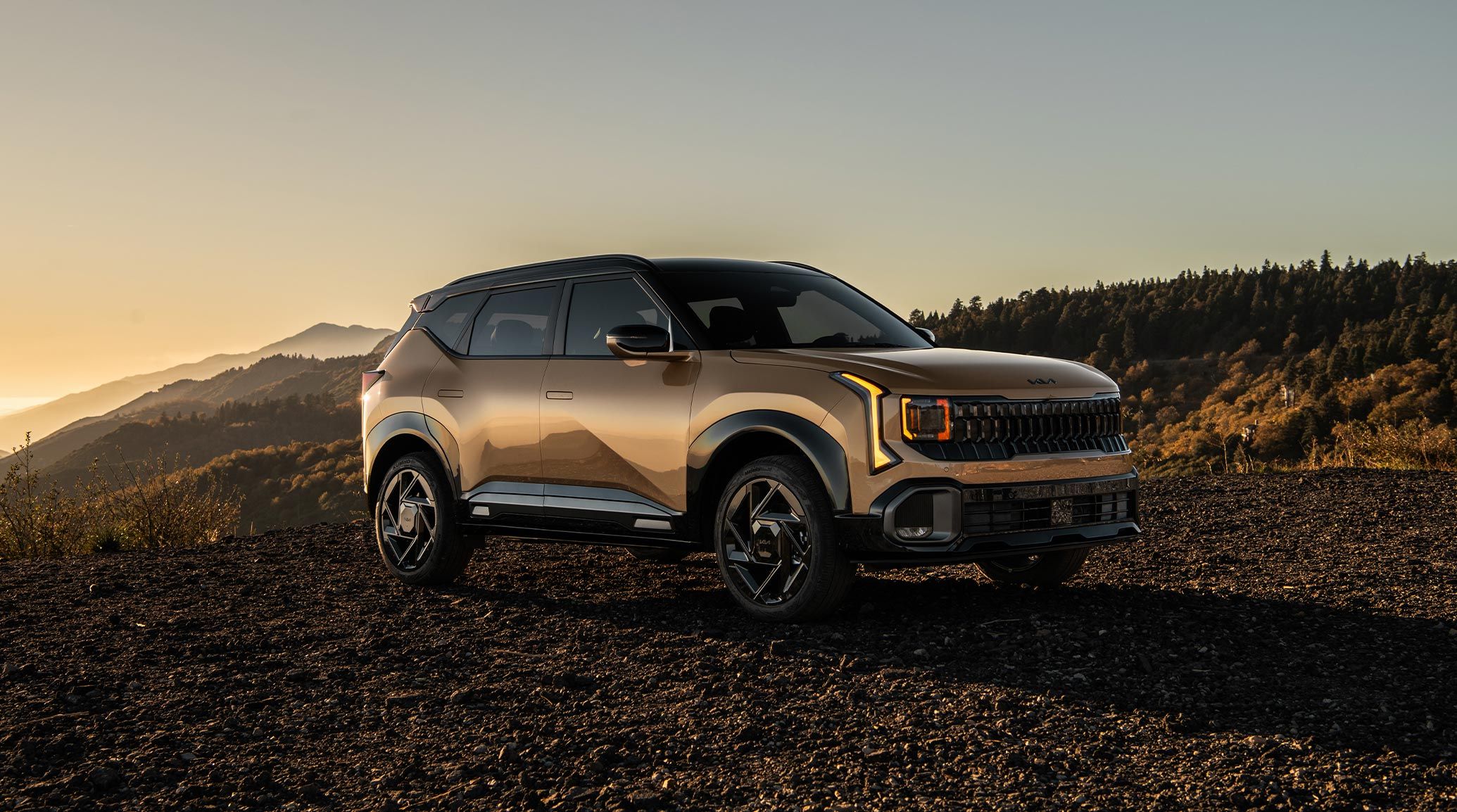 2027 Kia Seltos in brown parked outdoors at sunset, shown in a front three-quarter view with gloss black exterior trim, black side mirrors, and rugged styling against a mountain landscape.